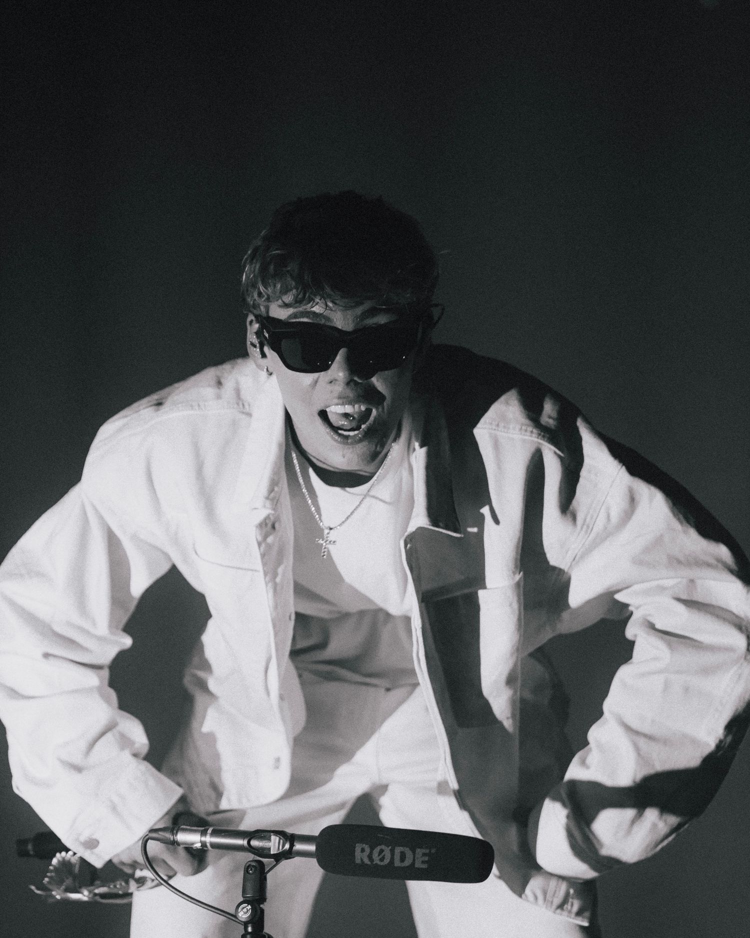 Black and white photo of someone in a white jacket and sunglasses posing with a microphone in a dramatic pose.