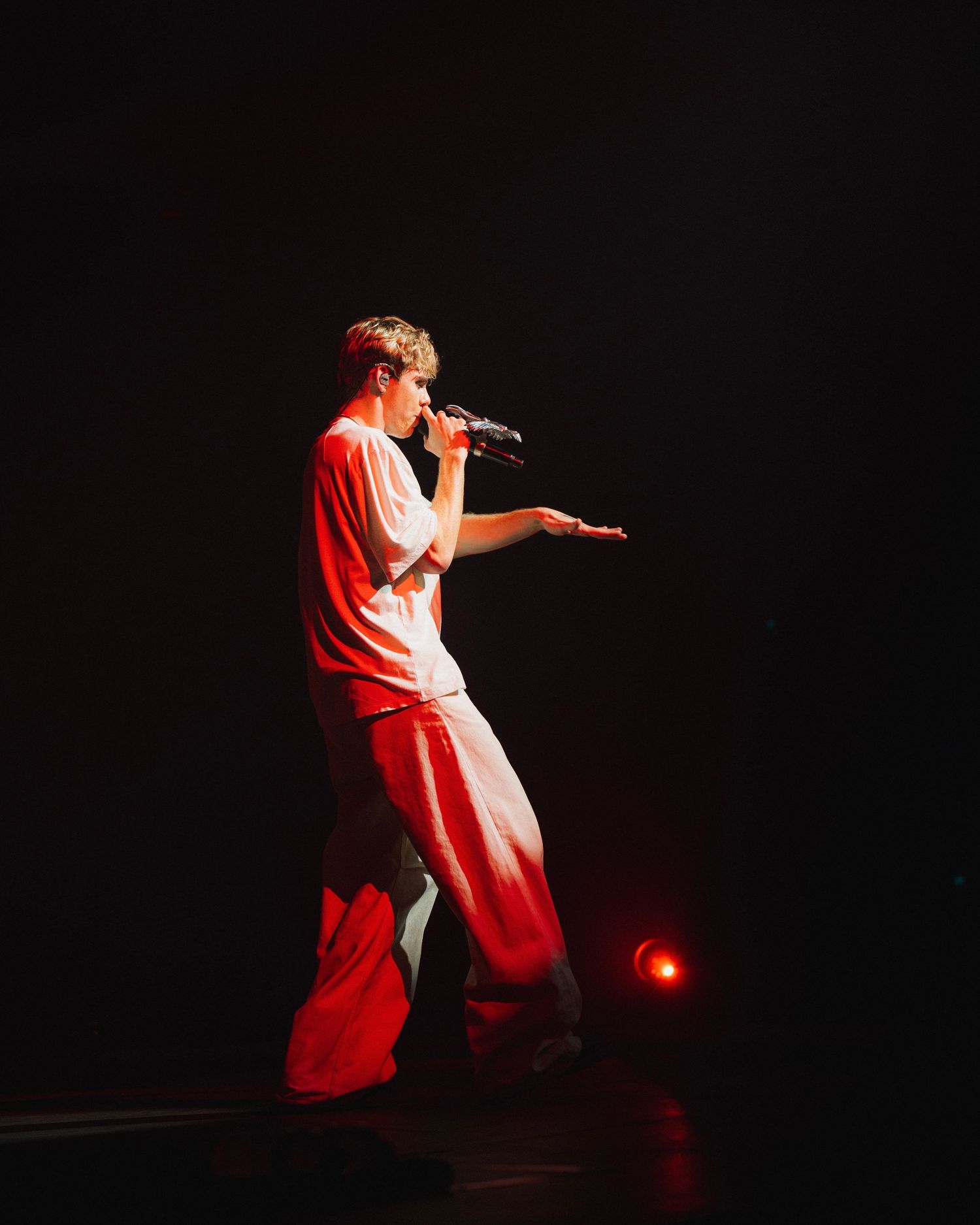 Performer in red stage lighting sings into microphone and gestures dramatically against dark background during concert.