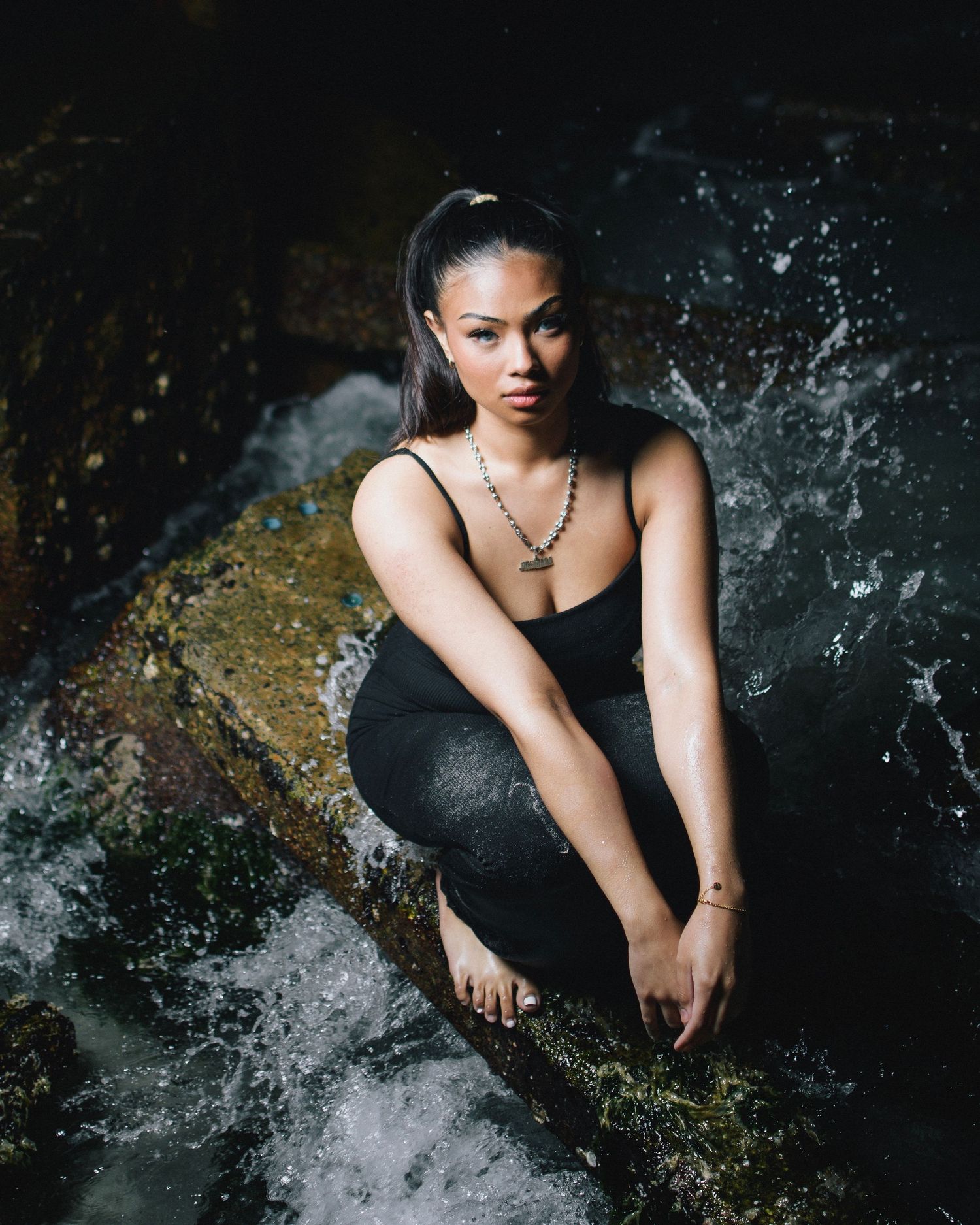 Dark artistic fashion photo of someone in black attire crouching near splashing water against a moody backdrop.
