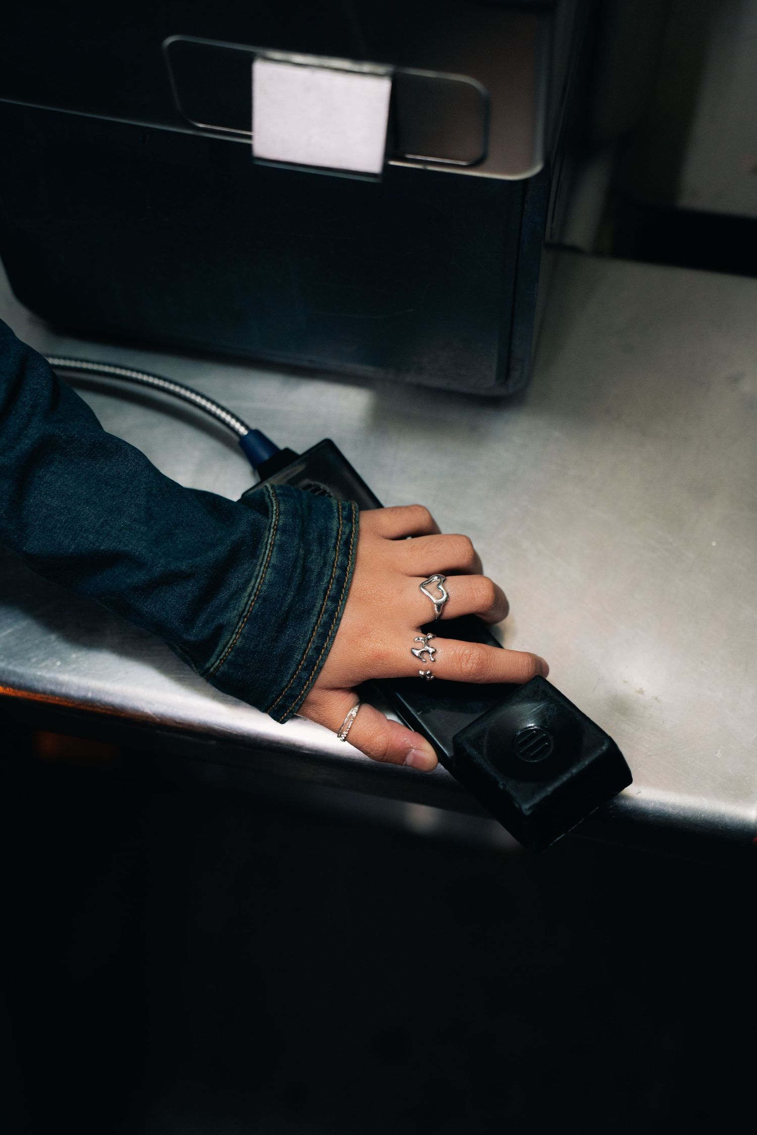 Hand wearing decorative rings and denim jacket sleeve resting on a dark surface in moody lighting.