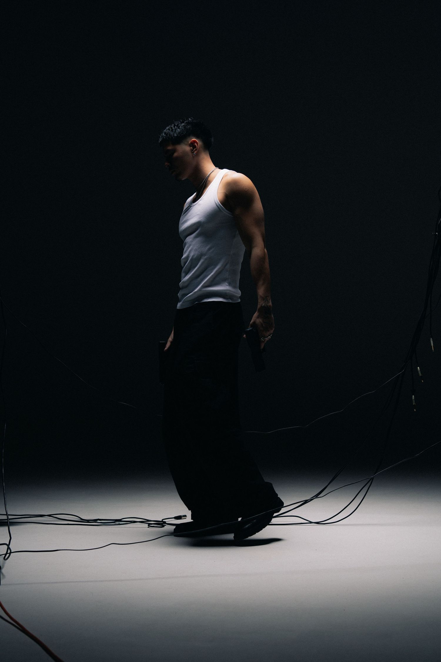A dramatic studio portrait of a person in a white tank top and black pants standing in shadow on a dark background.