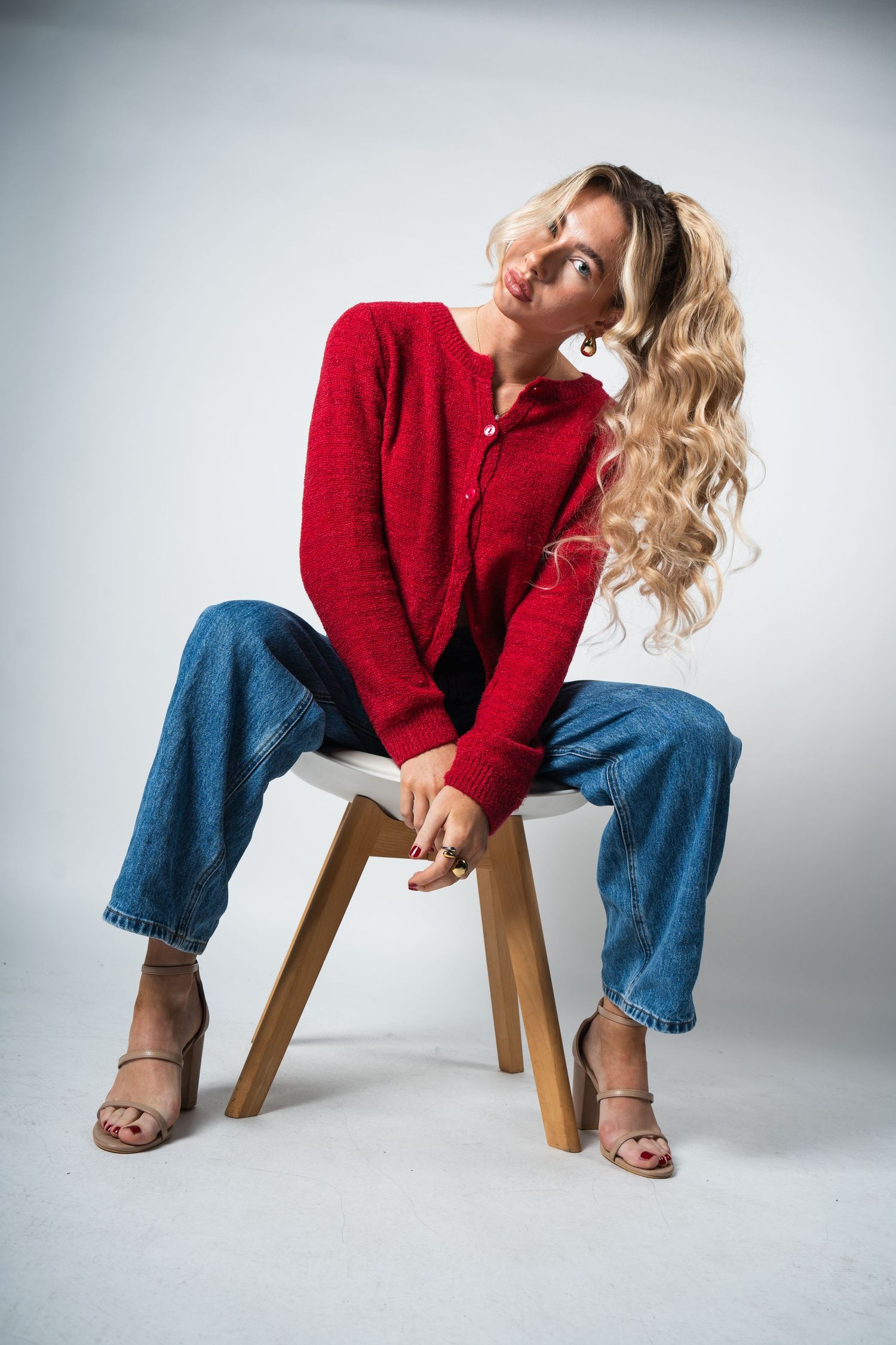 Model posing on wooden stool wearing red sweater, blue jeans and brown sandals against white background.