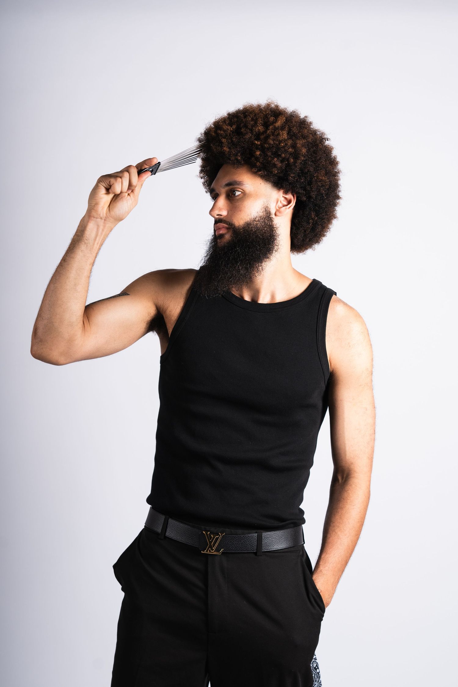 Person with an afro hairstyle and beard wearing black tank top and pants poses with a comb against light background.