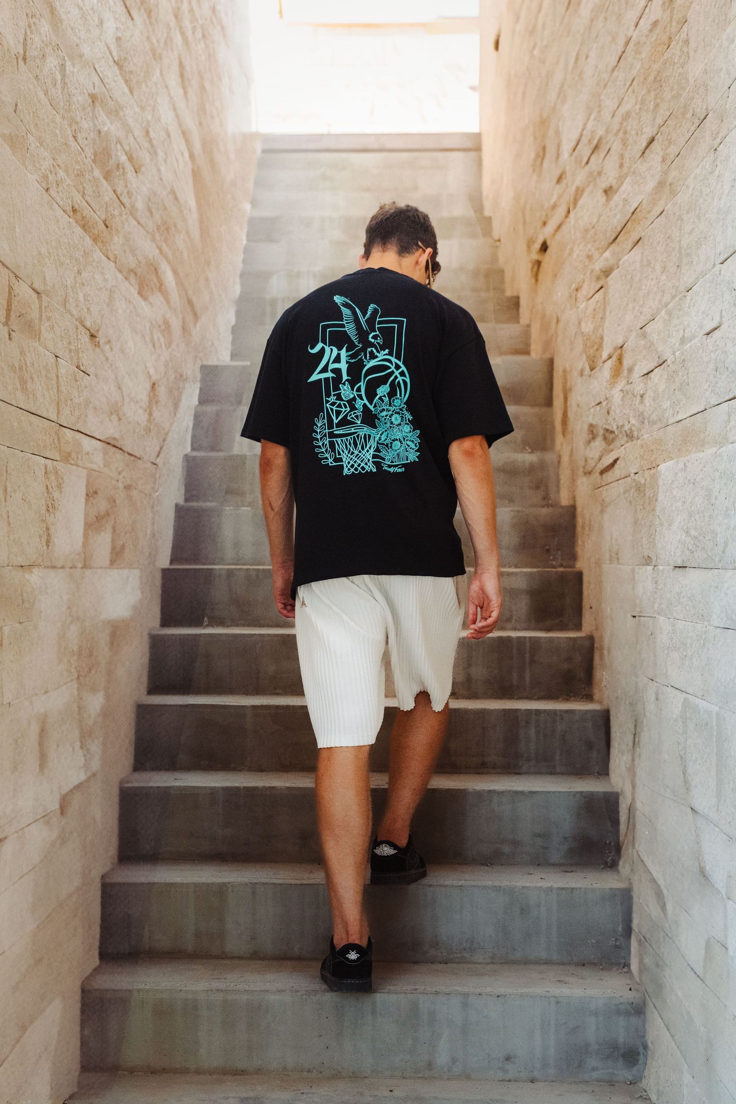 Person wearing black graphic t-shirt and white shorts ascending stone staircase in narrow passageway.