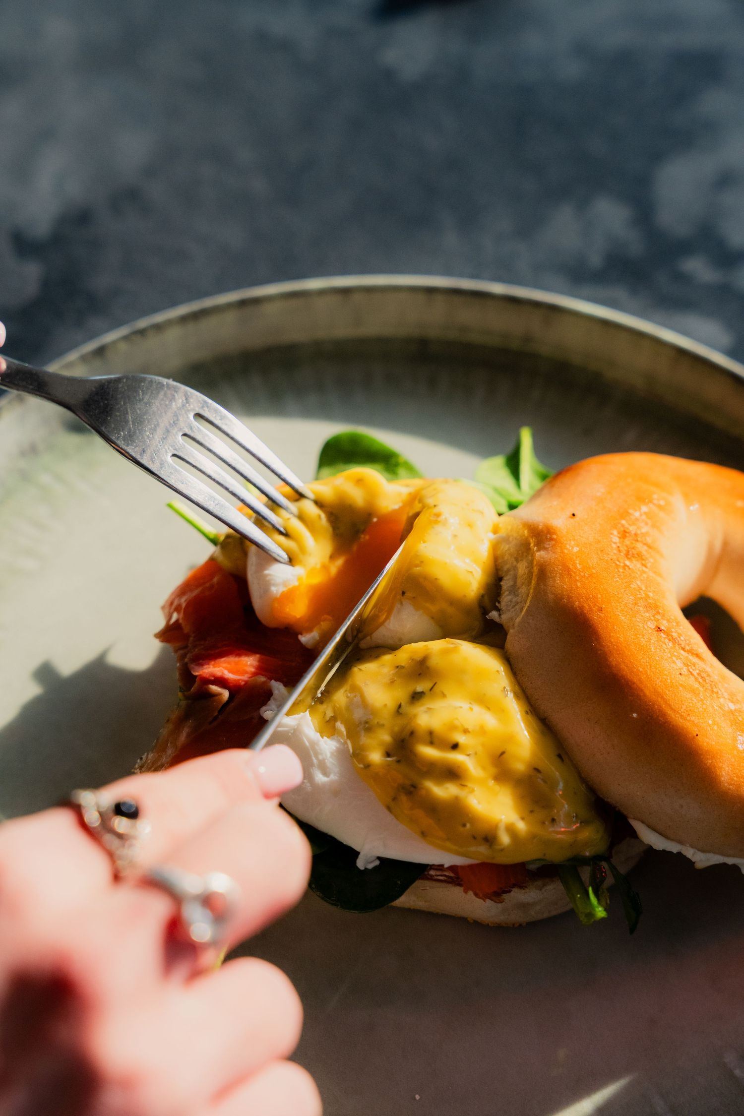 Hands cutting into a breakfast bagel topped with eggs and salmon.