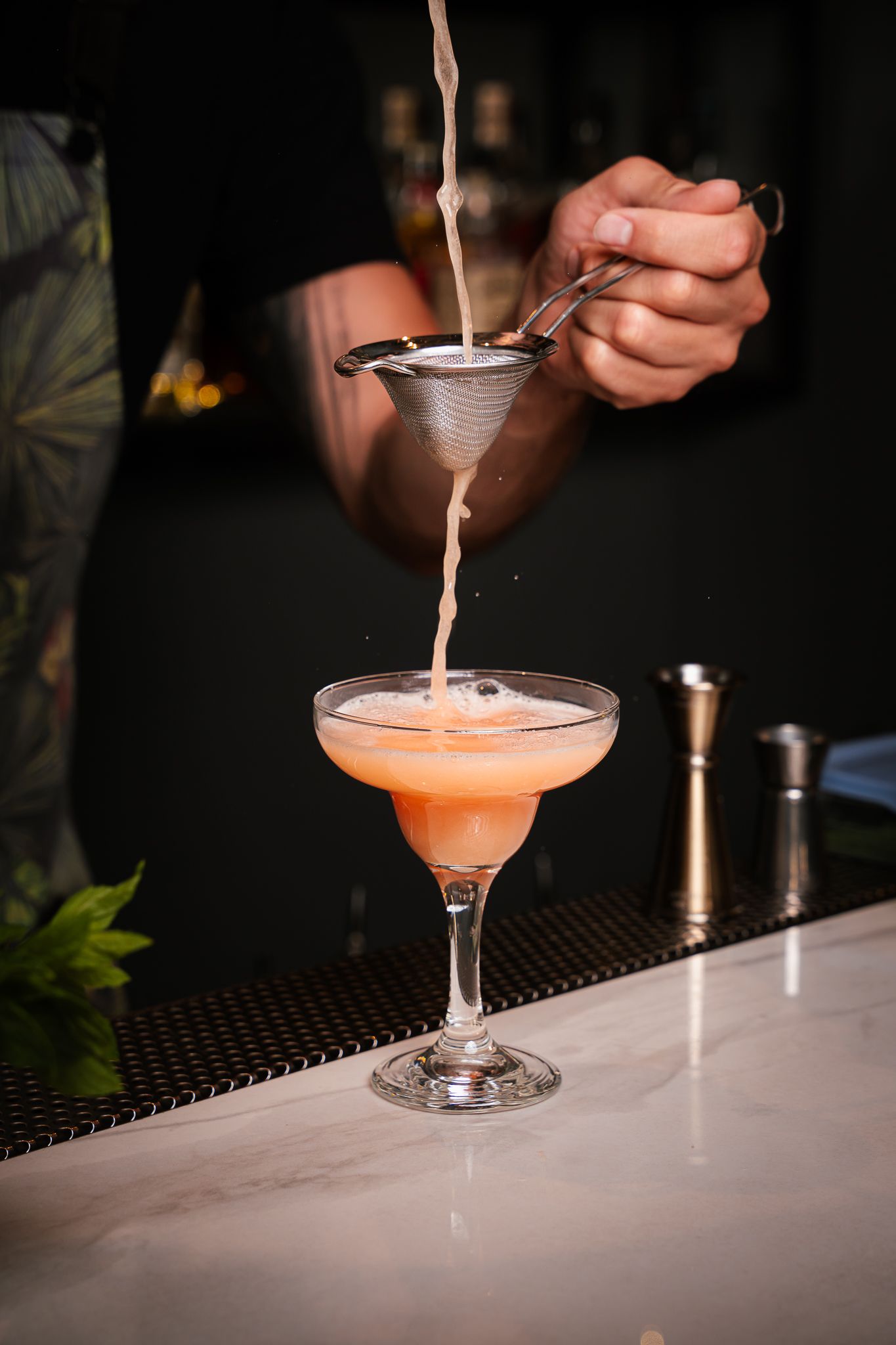 Pink cocktail being poured into coupe glass on bar counter with dark background.