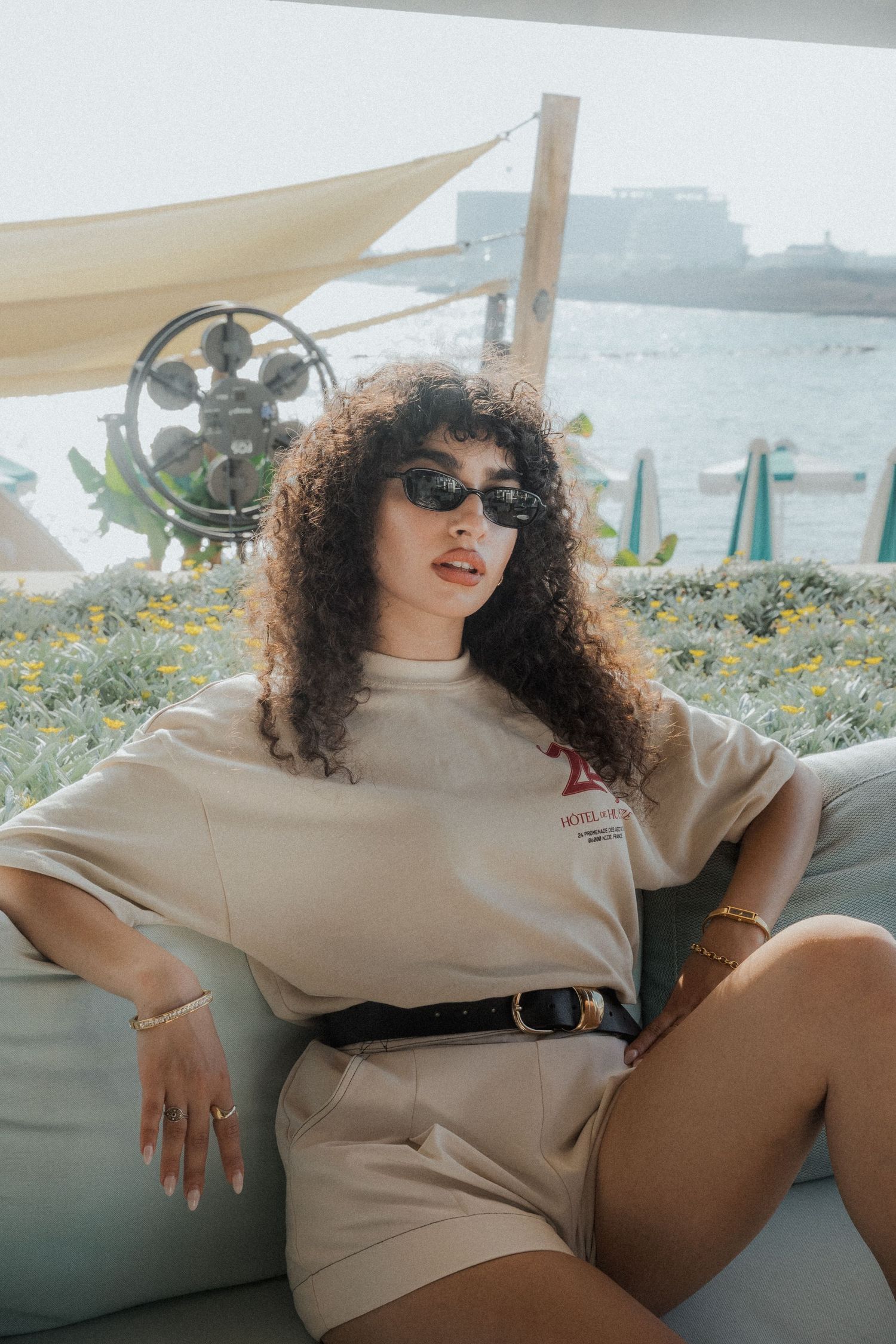 Woman with curly hair wearing beige outfit lounging on outdoor patio furniture.