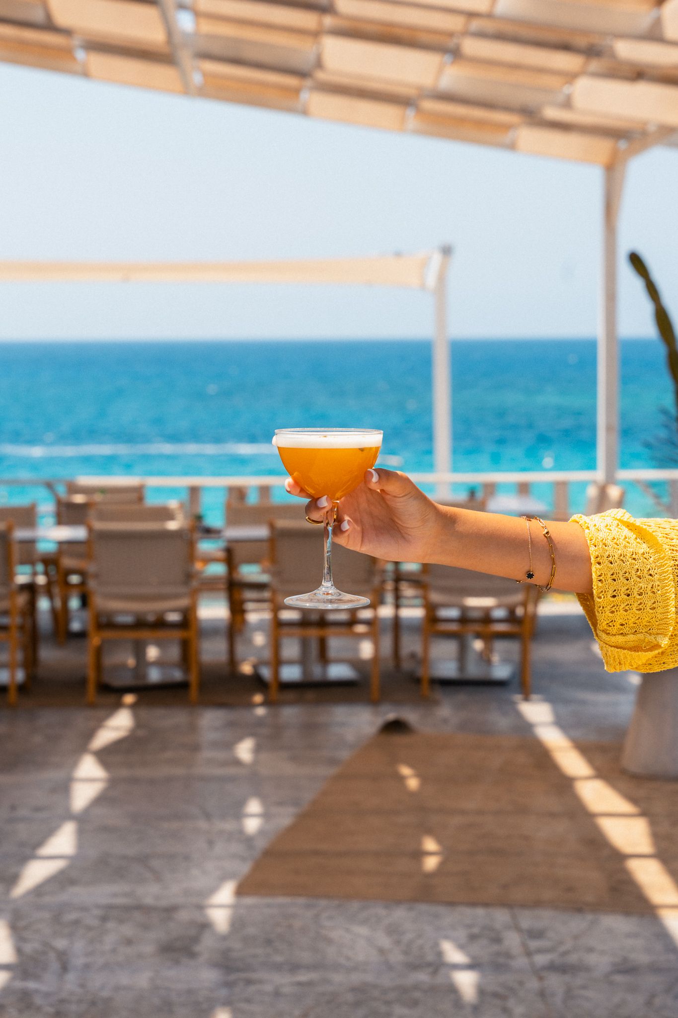 Beachside restaurant terrace with orange cocktails and wooden furniture overlooking turquoise ocean waters.