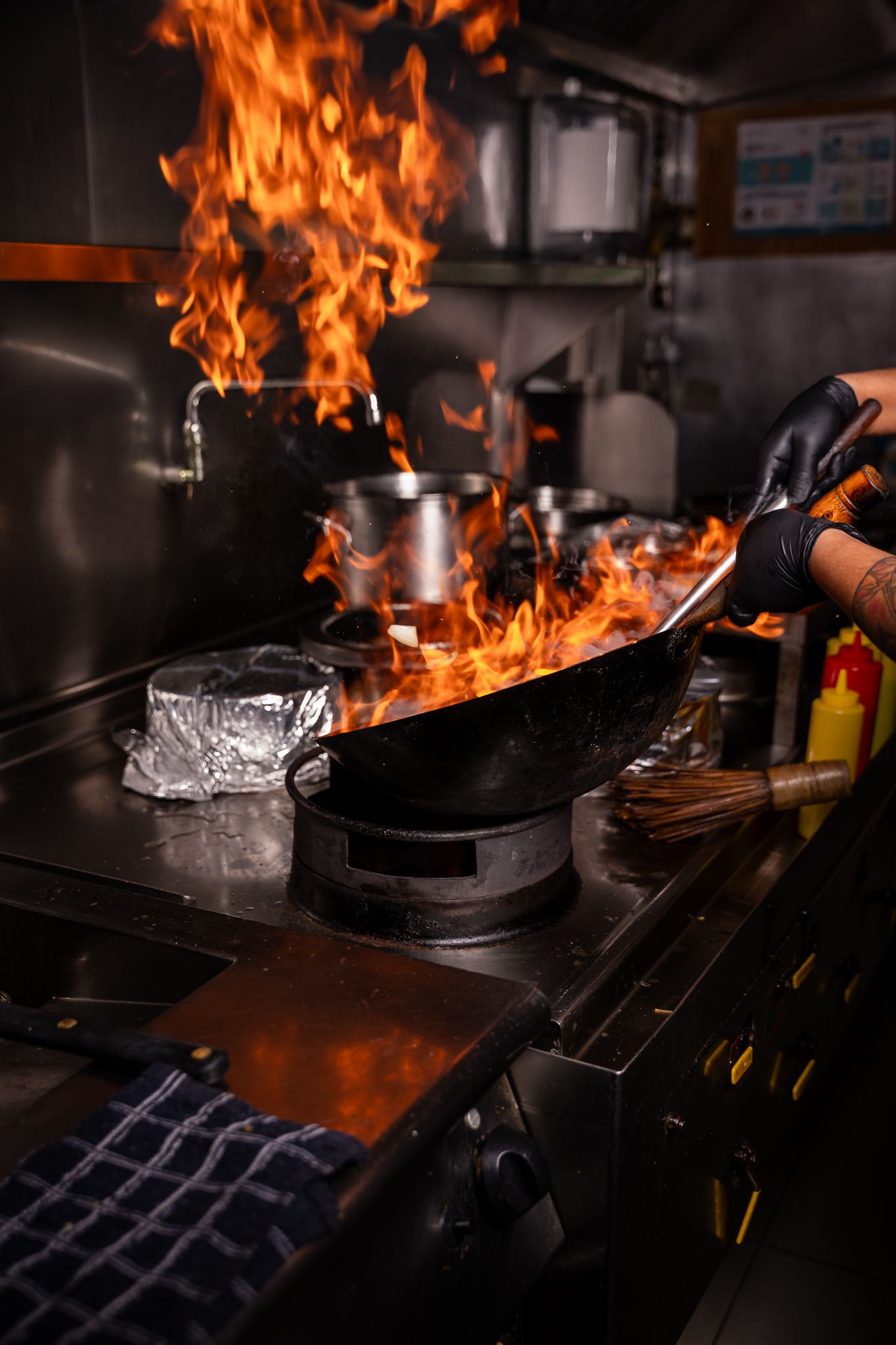 Professional chef cooking with intense flames in a dark commercial kitchen with multiple fiery wok pans on the stove.