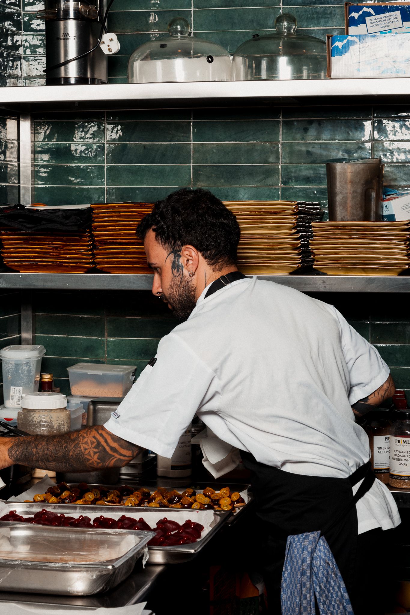 Chef in white coat reaches across kitchen counter to prepare food on stainless steel surfaces.