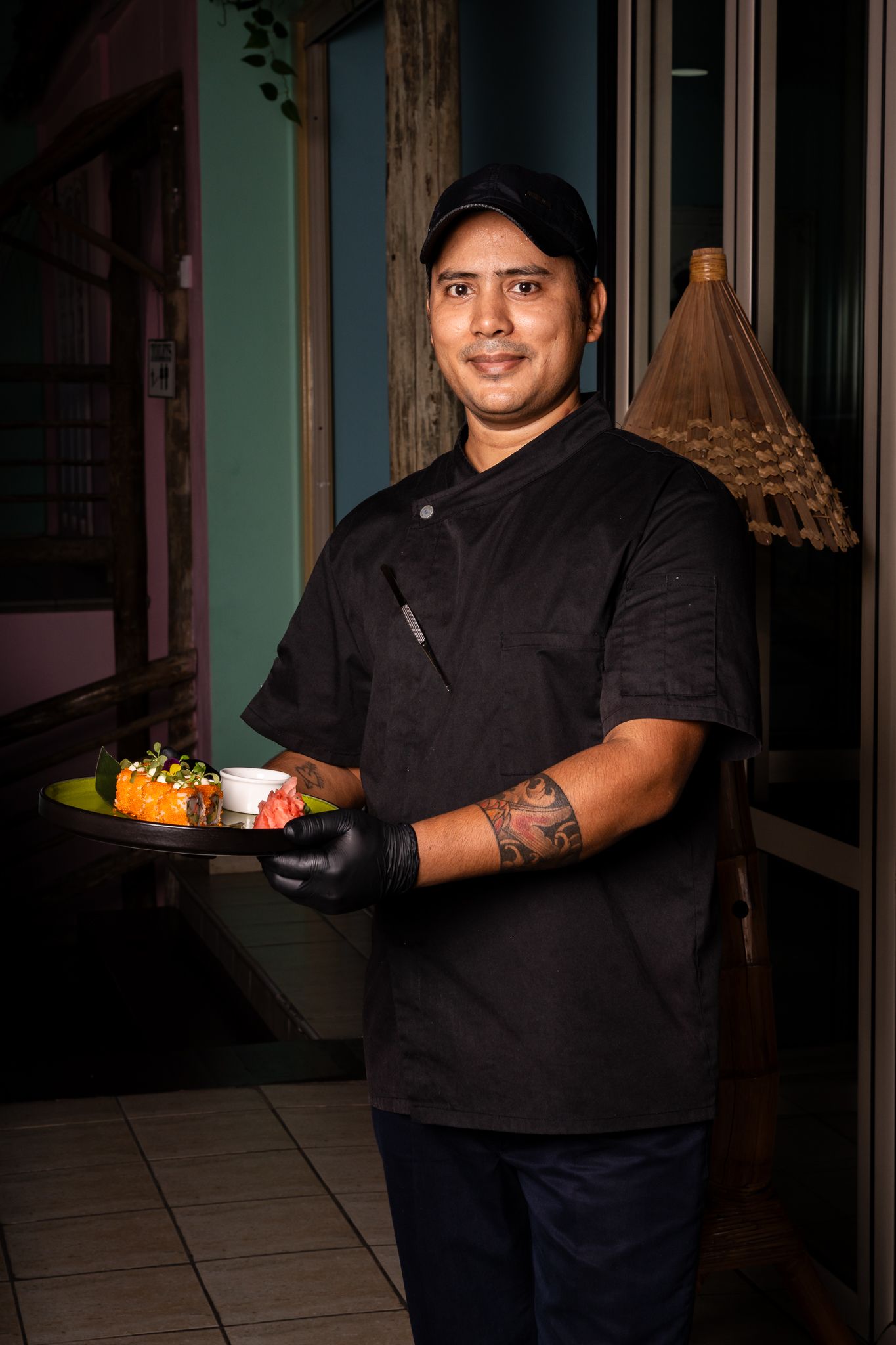 Restaurant worker in black uniform poses with plate of food in dimly lit dining room.