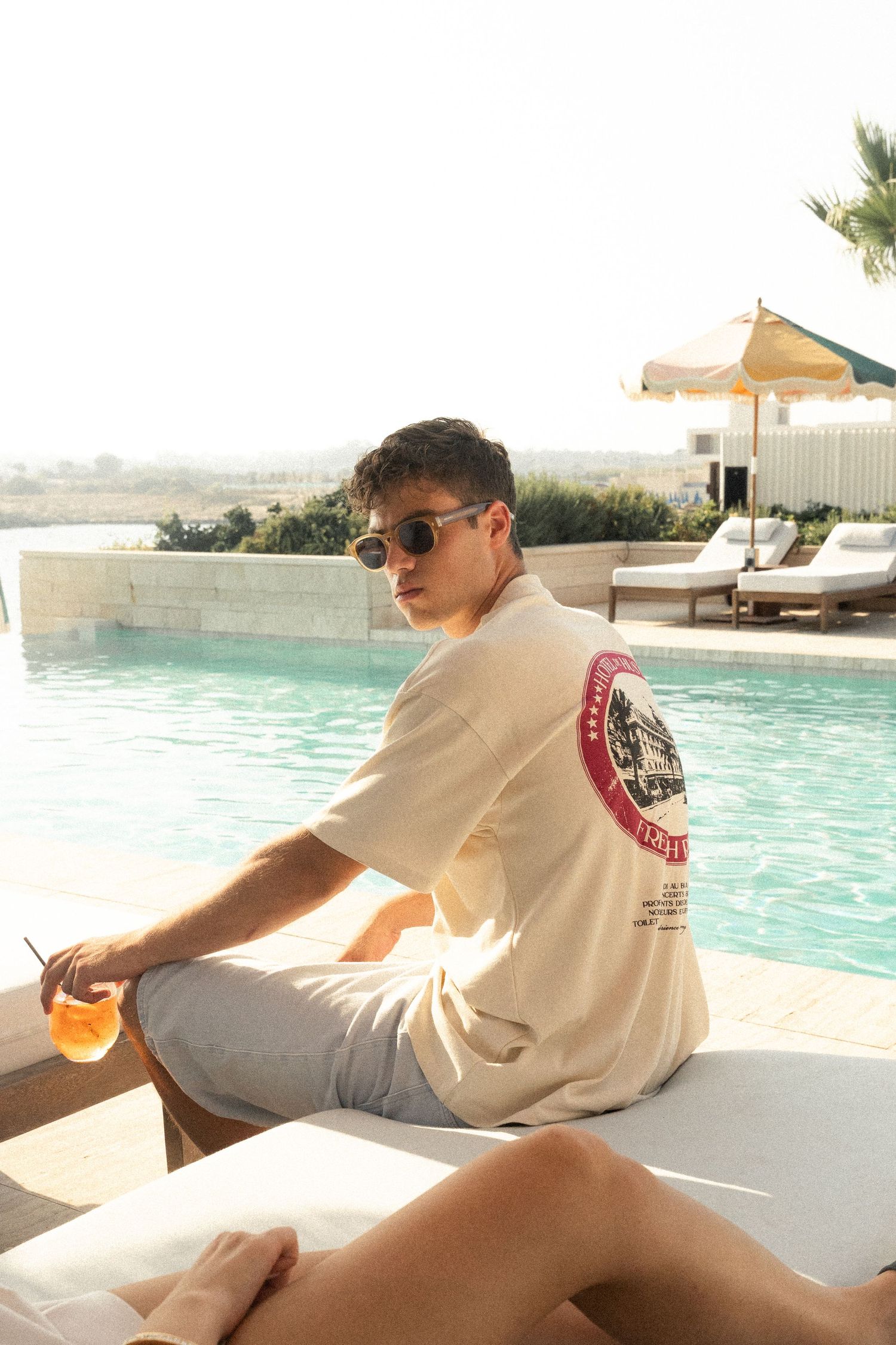 Person in beige t-shirt relaxing poolside with orange drink at luxury resort.