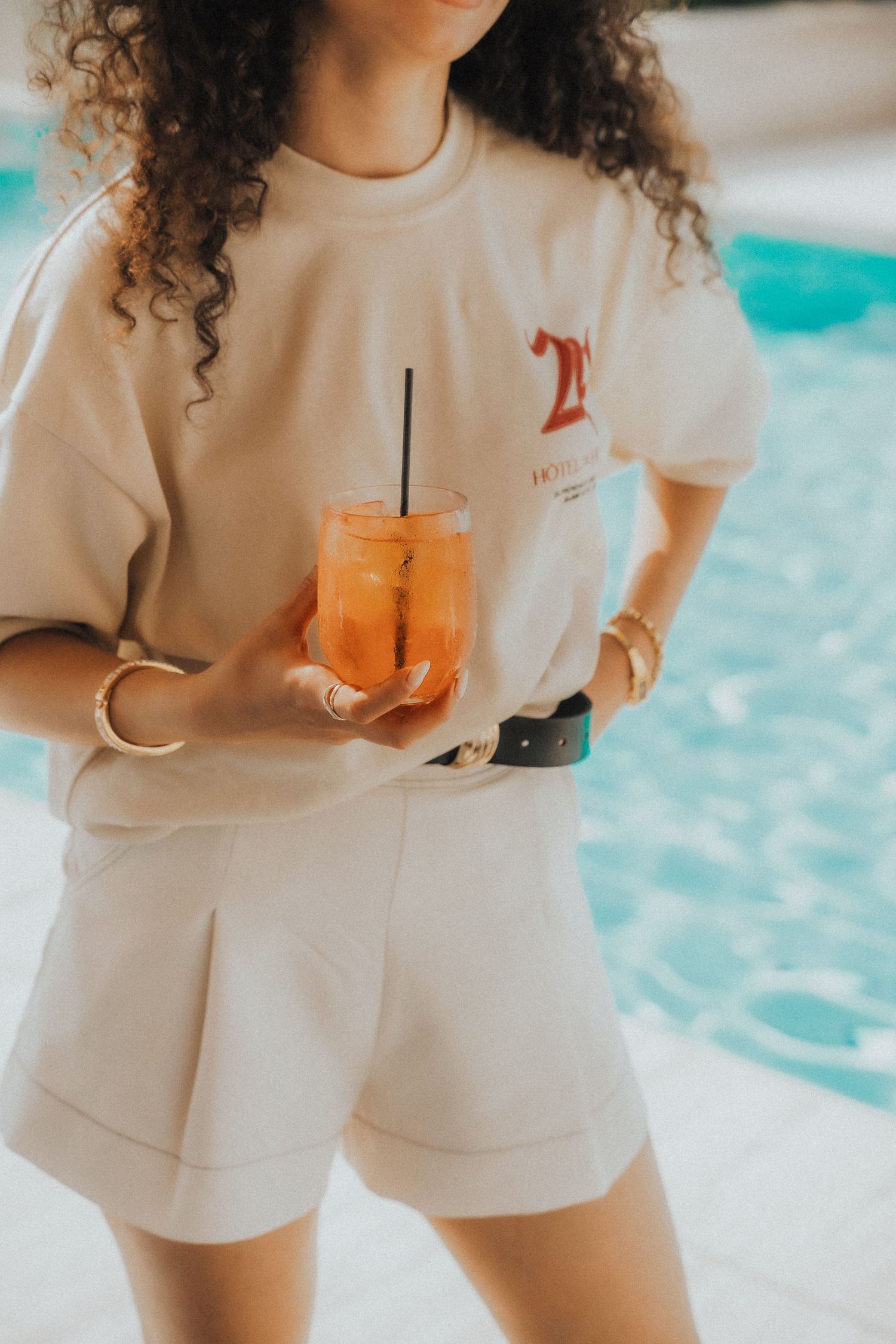Person in white sweatshirt and shorts holding orange tropical drink by poolside.