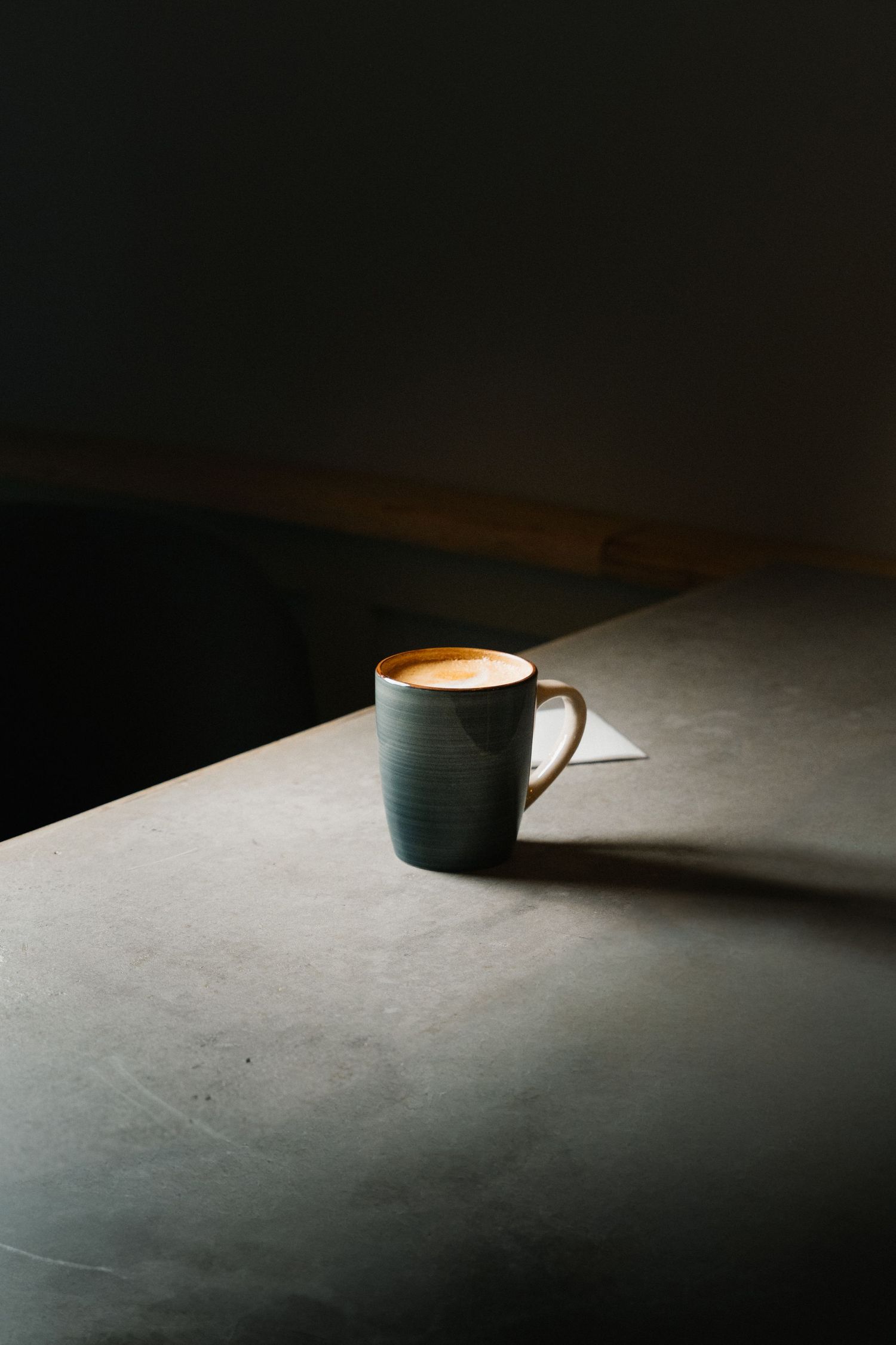 A dark green ceramic mug with latte art sits alone on a gray surface in moody lighting.