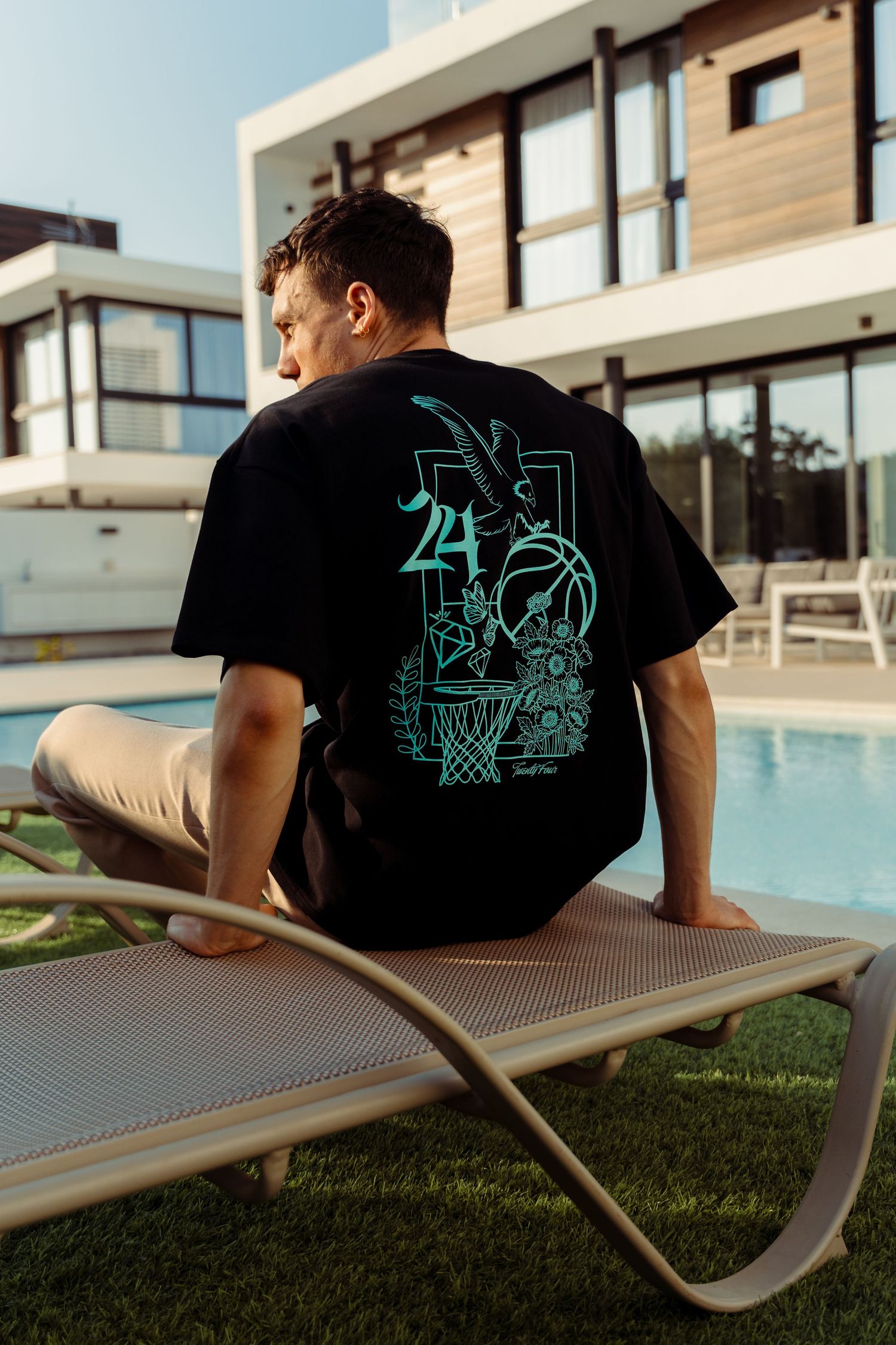 Man in black graphic t-shirt sitting on lounge chair by modern poolside.