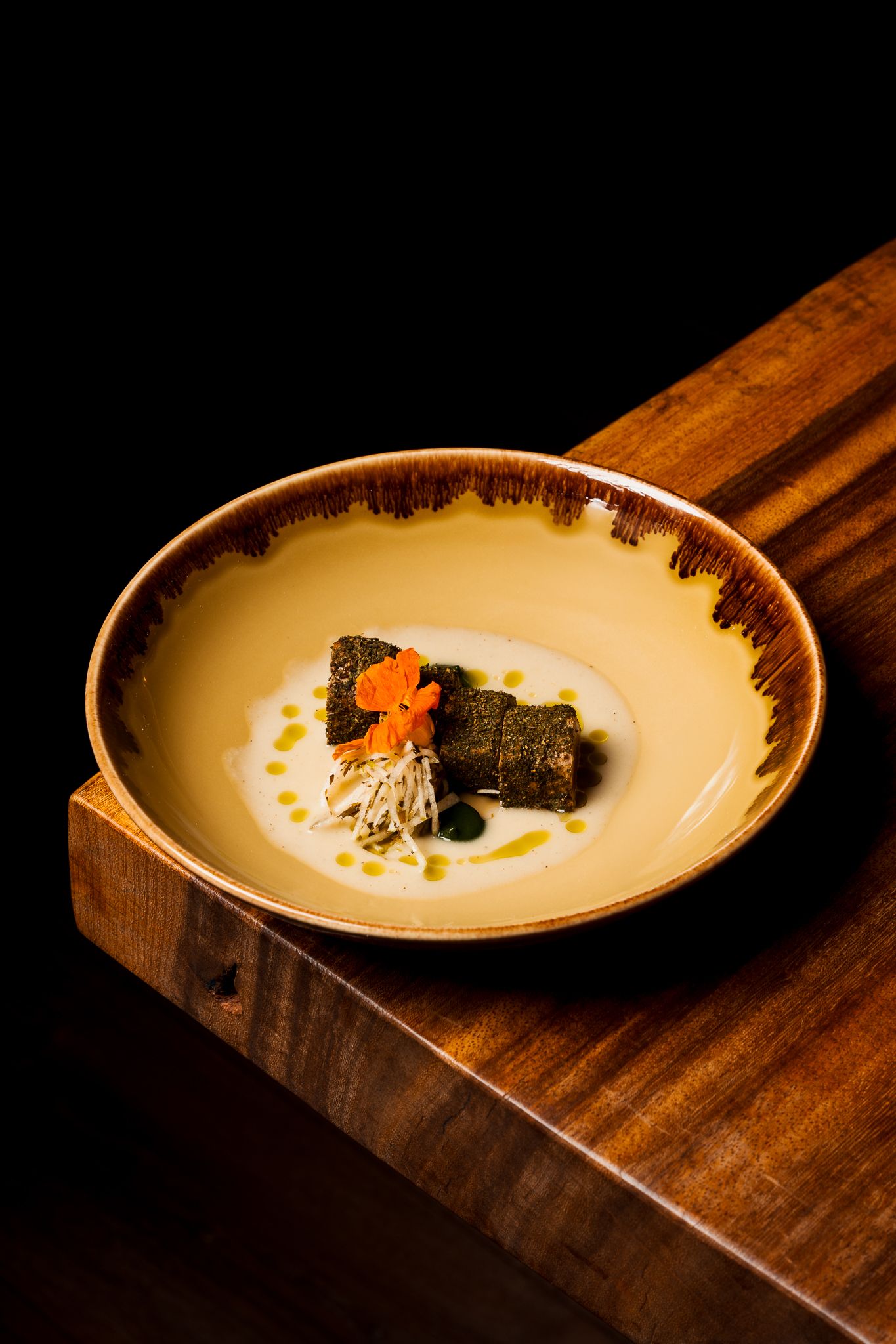 A gourmet plated dish featuring dark garnish and orange accent on a cream-colored ceramic bowl with brown rim.