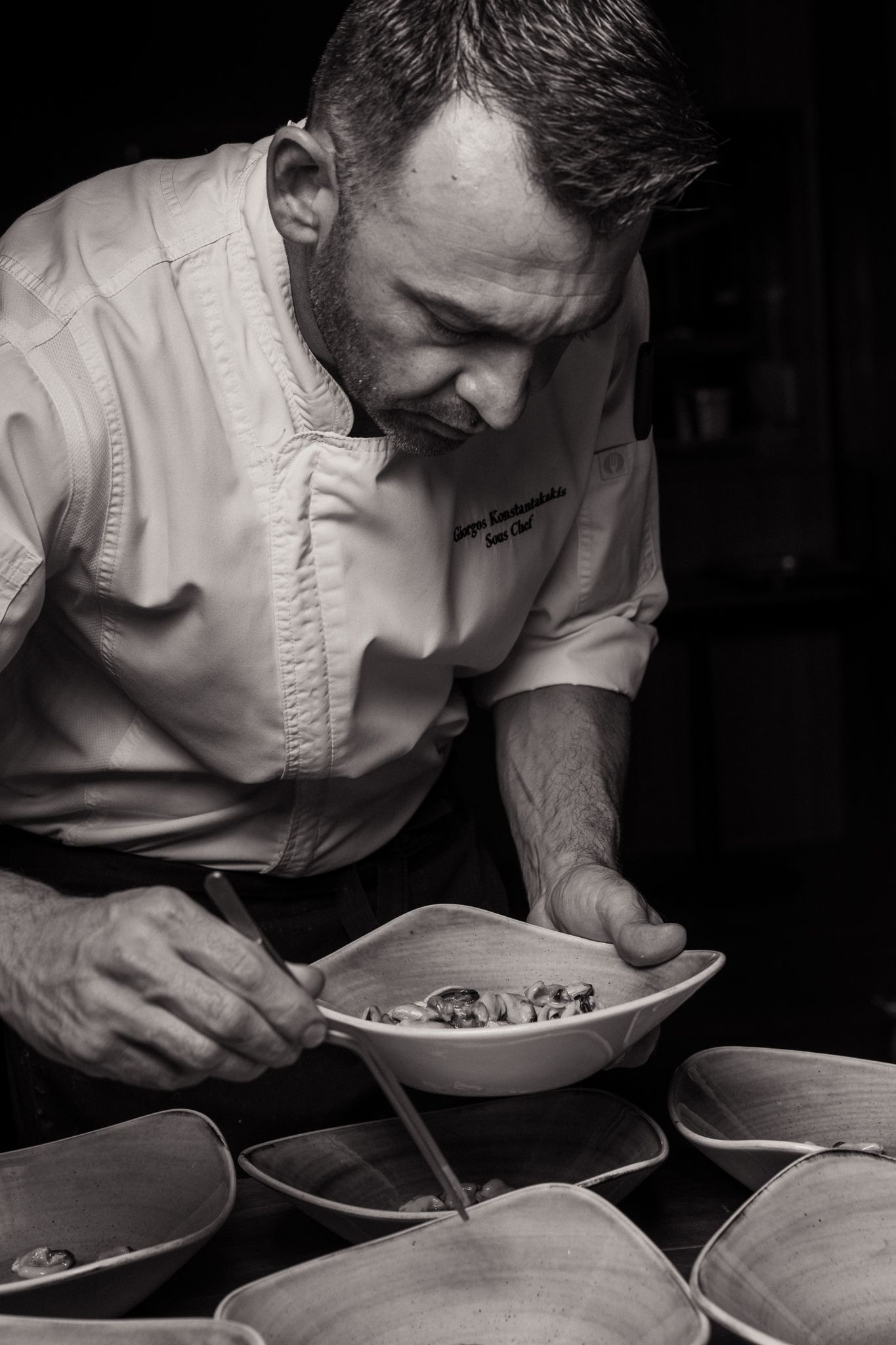 Chef closely inspects plated dishes in restaurant kitchen shown in dramatic black and white photography series.