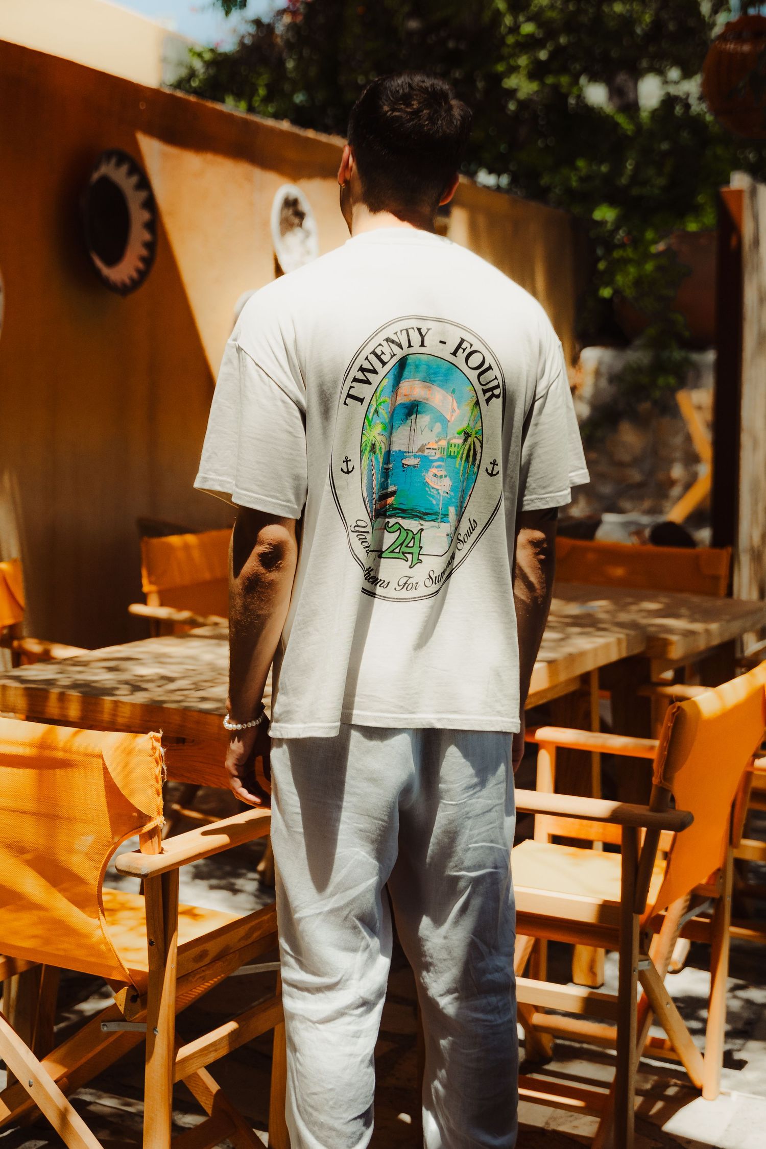 Person wearing white t-shirt with beach graphic design standing among wooden chairs outdoors.