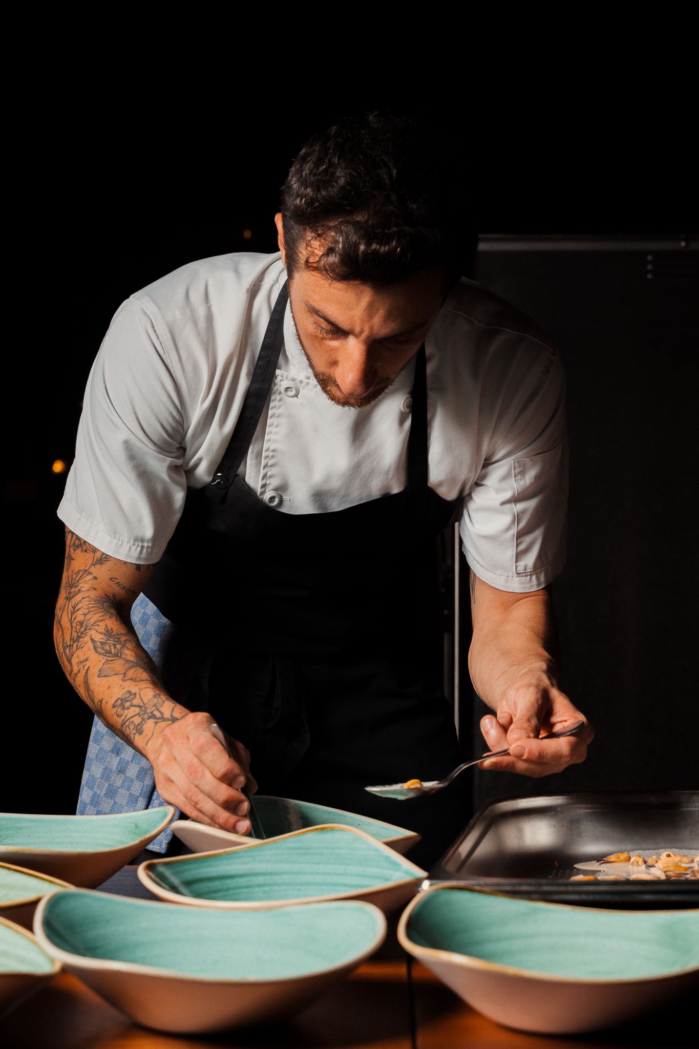 Chef in white uniform carefully plates a dish on turquoise ceramic plates in a dark professional kitchen setting.