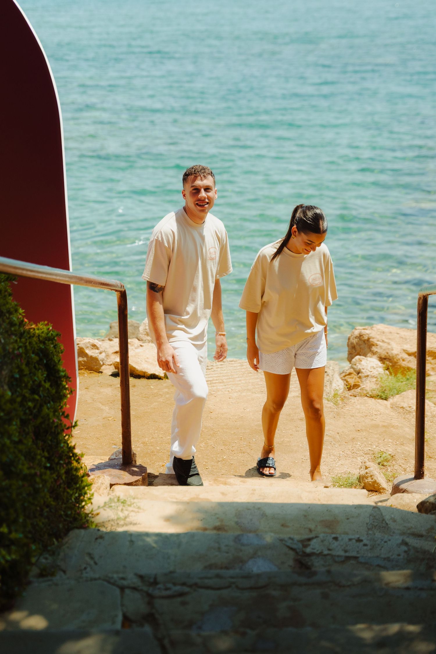 Two people in light colored clothing standing on coastal pathway overlooking turquoise water.