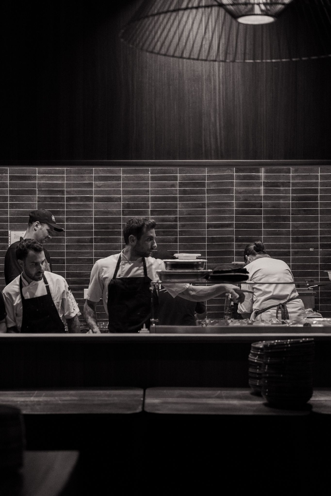 Chefs working together in a modern restaurant kitchen with tiled walls and dramatic lighting.