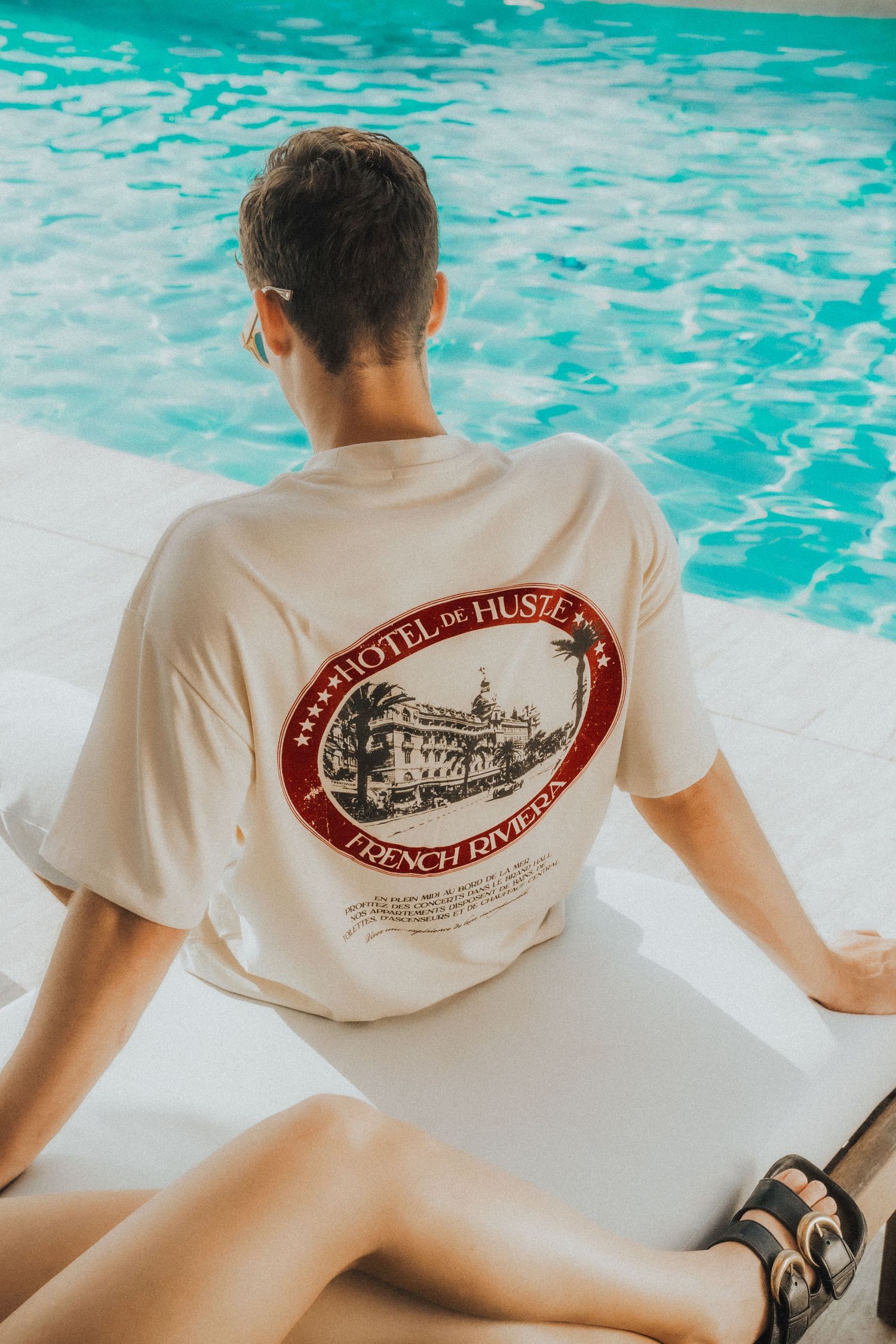 Person wearing white t-shirt with vintage hotel logo relaxing by turquoise pool.