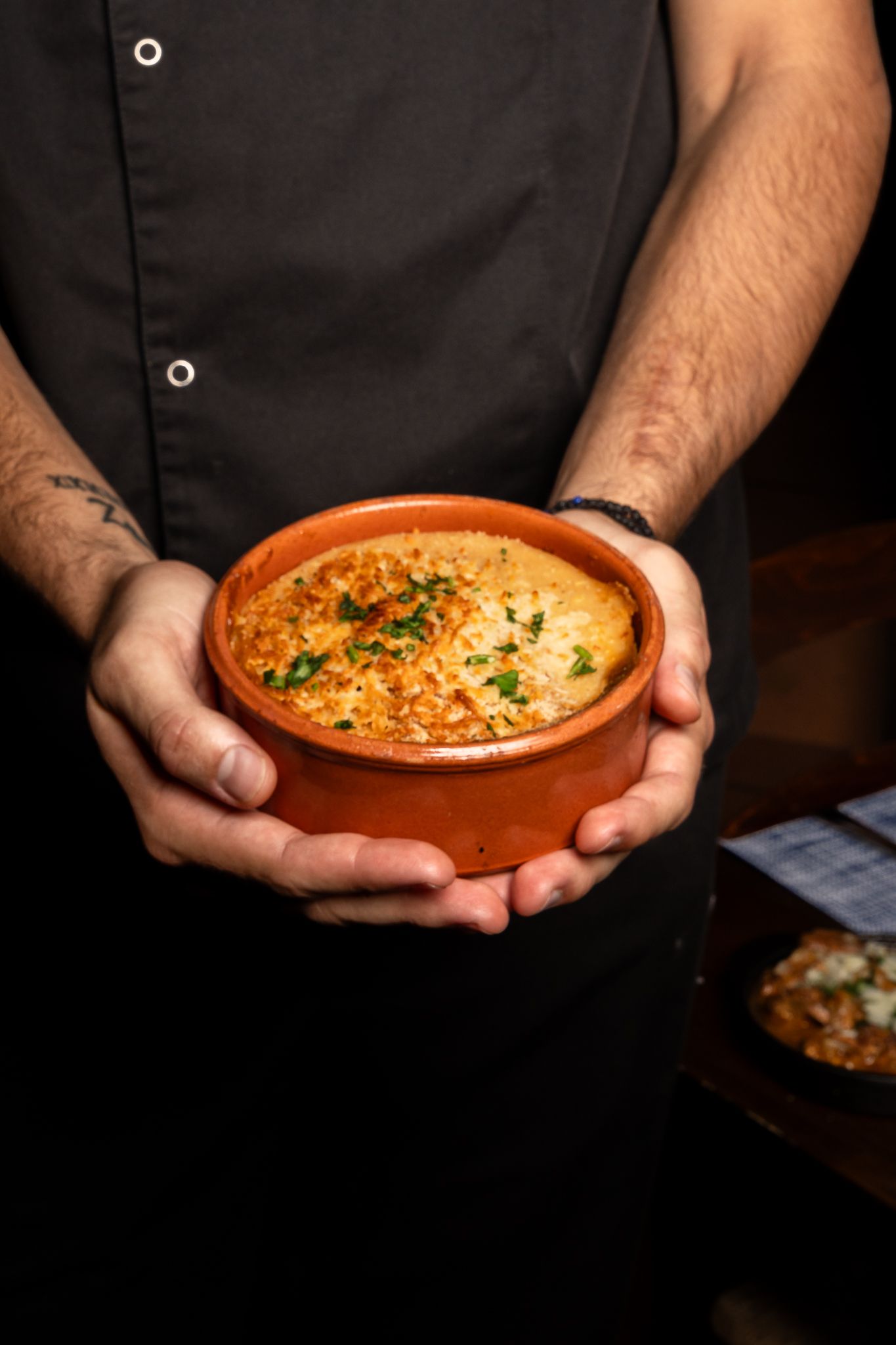 Chef holding a rustic clay bowl of creamy baked dish garnished with herbs.