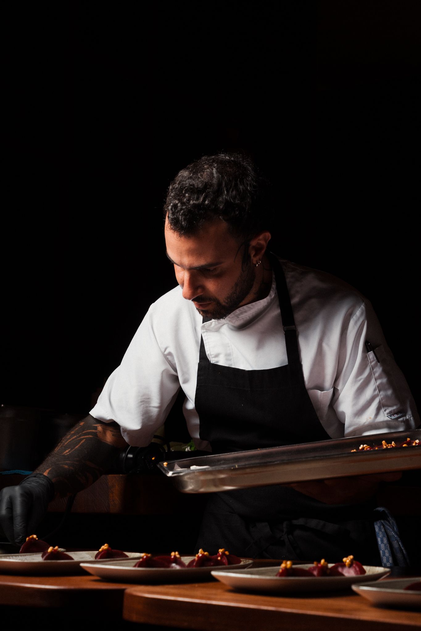 Chef in white uniform plating food in dark moody lighting at professional kitchen counter.