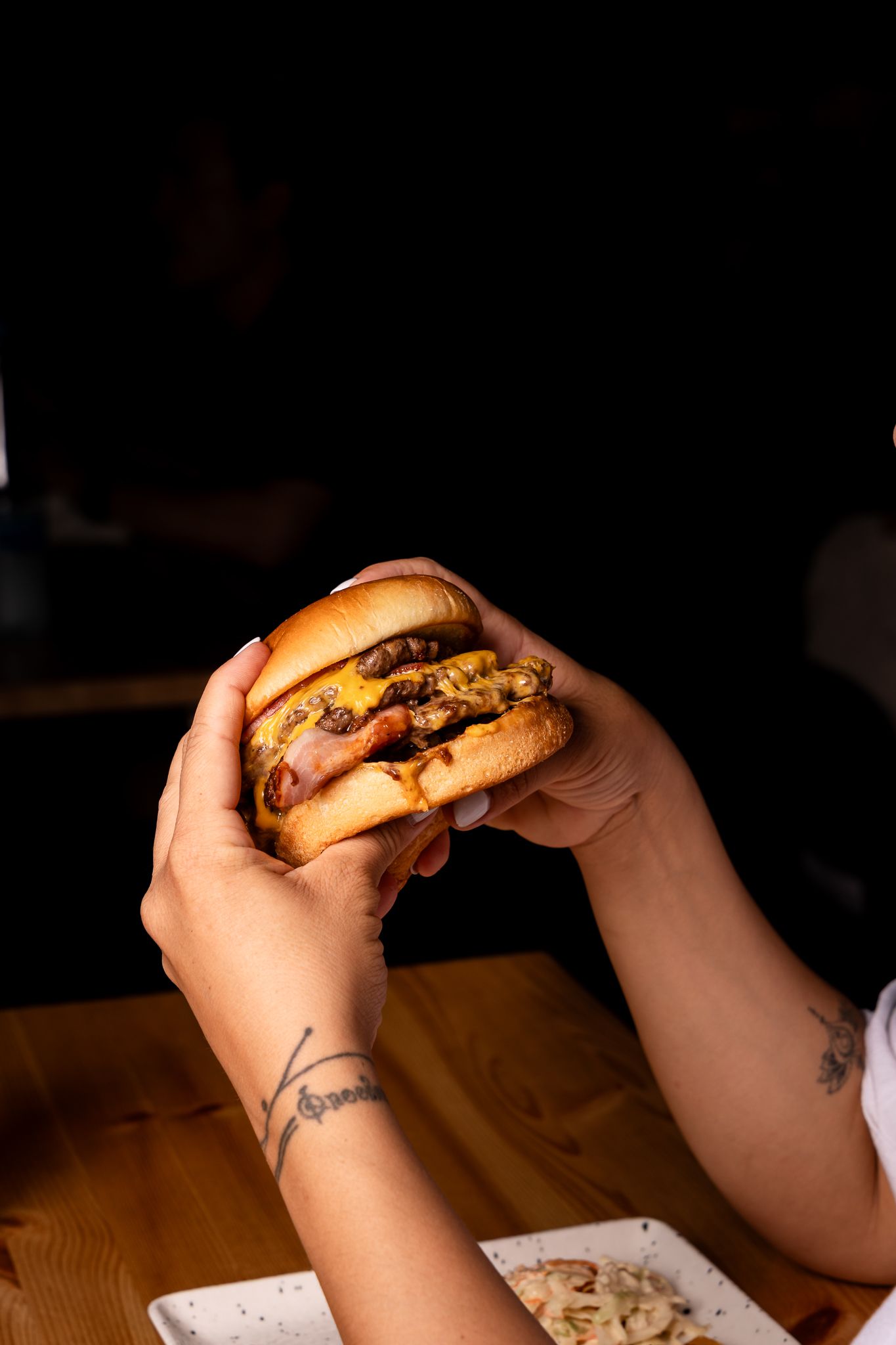 Hands holding and preparing to eat a juicy cheeseburger in dim lighting.