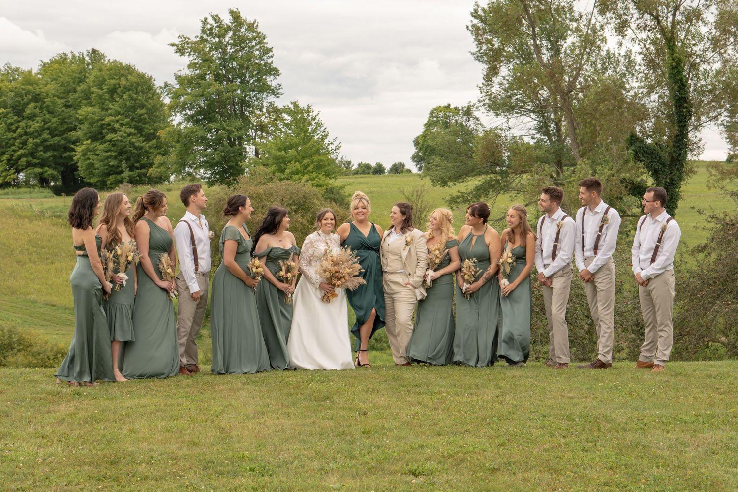 Wedding party in sage green dresses and khaki suits posing together on a grassy hillside.