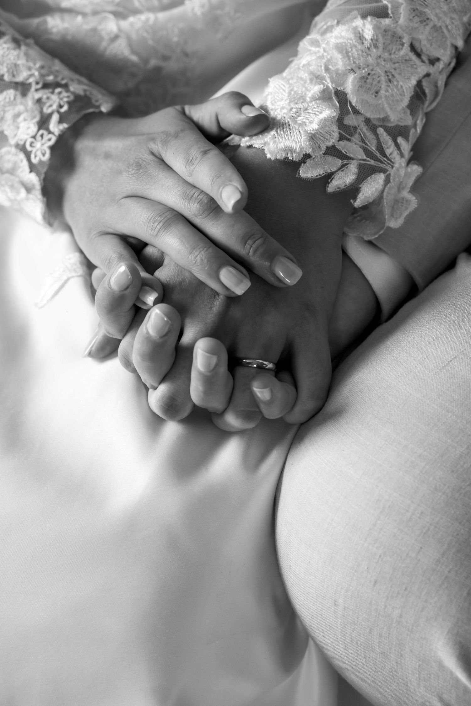 Black and white close-up of hands with wedding rings and lace dress details.