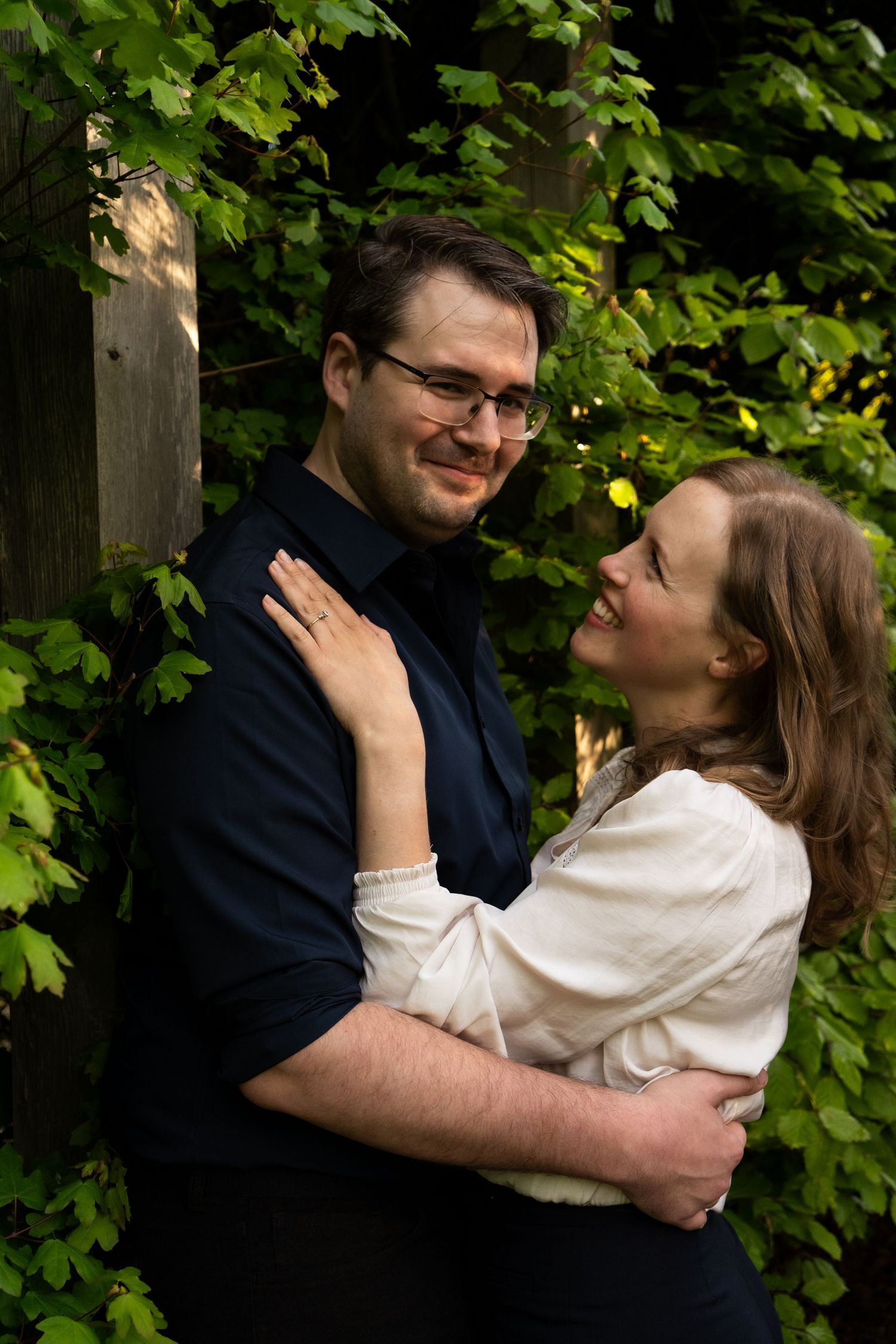 A romantic couple embraces in front of green foliage during an outdoor photo session.