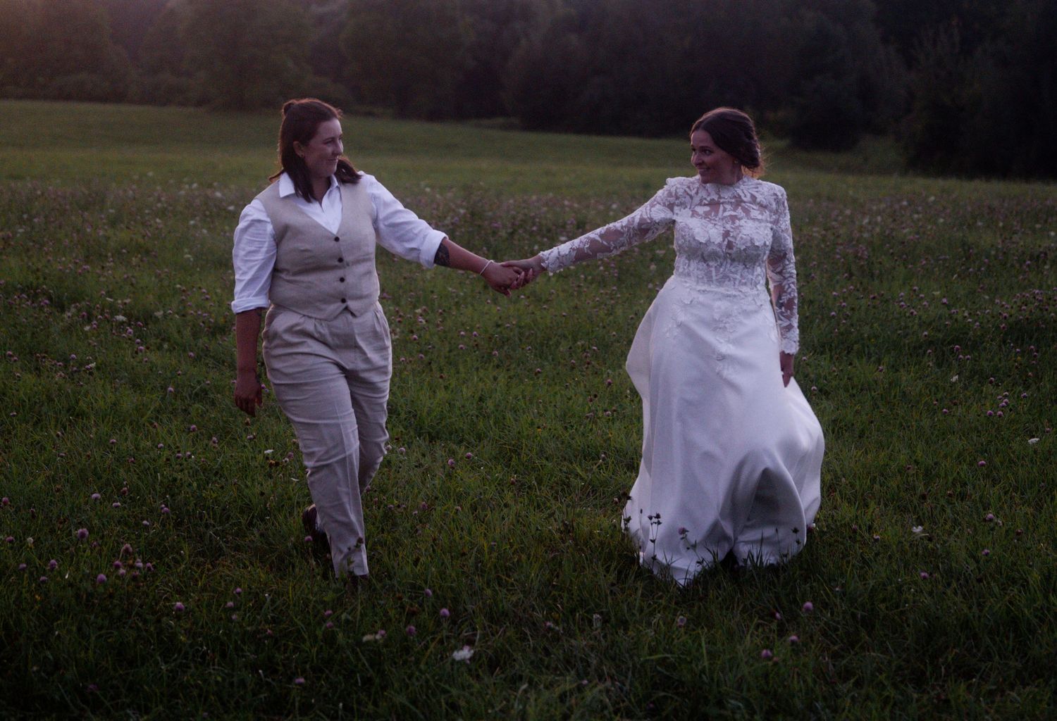 Couple holds hands while walking through a dusky meadow in wedding attire.