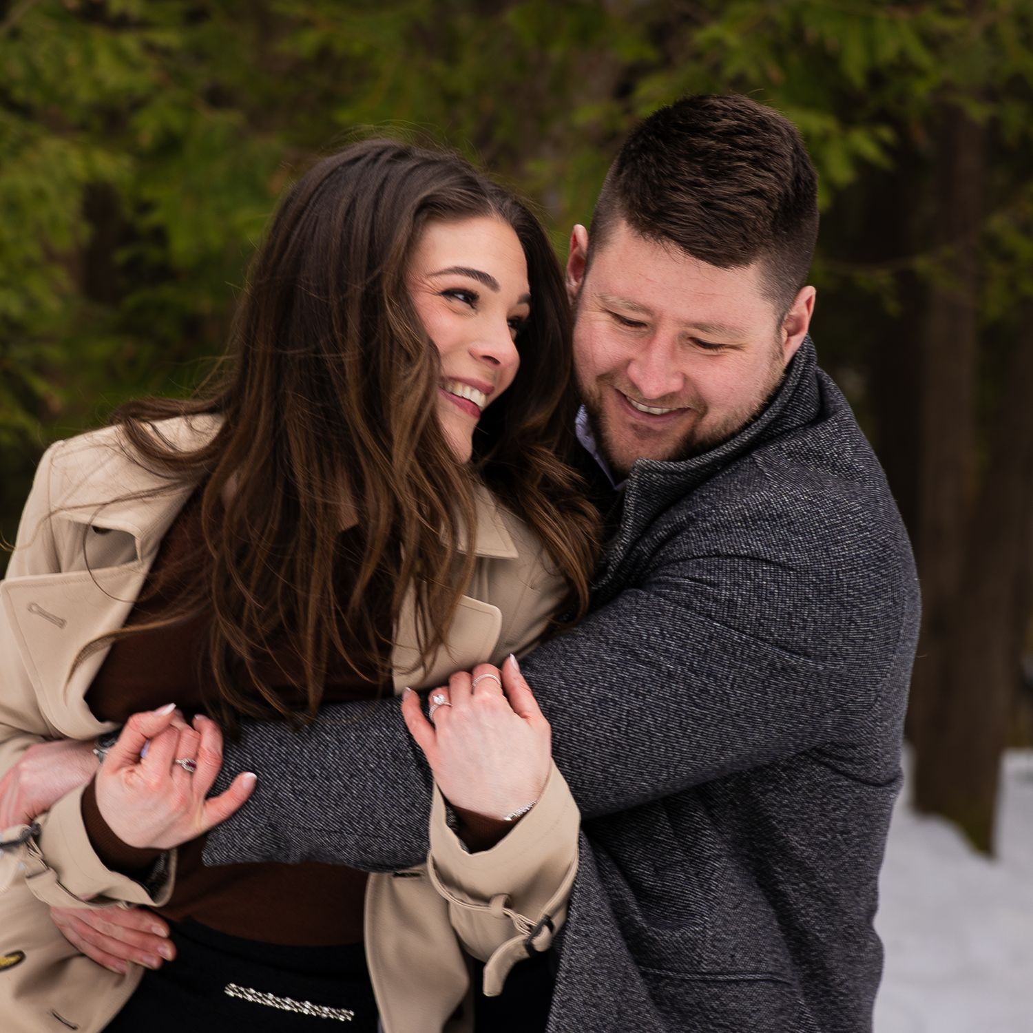A couple laughs and embraces while walking through a snowy winter landscape in matching dark coats.