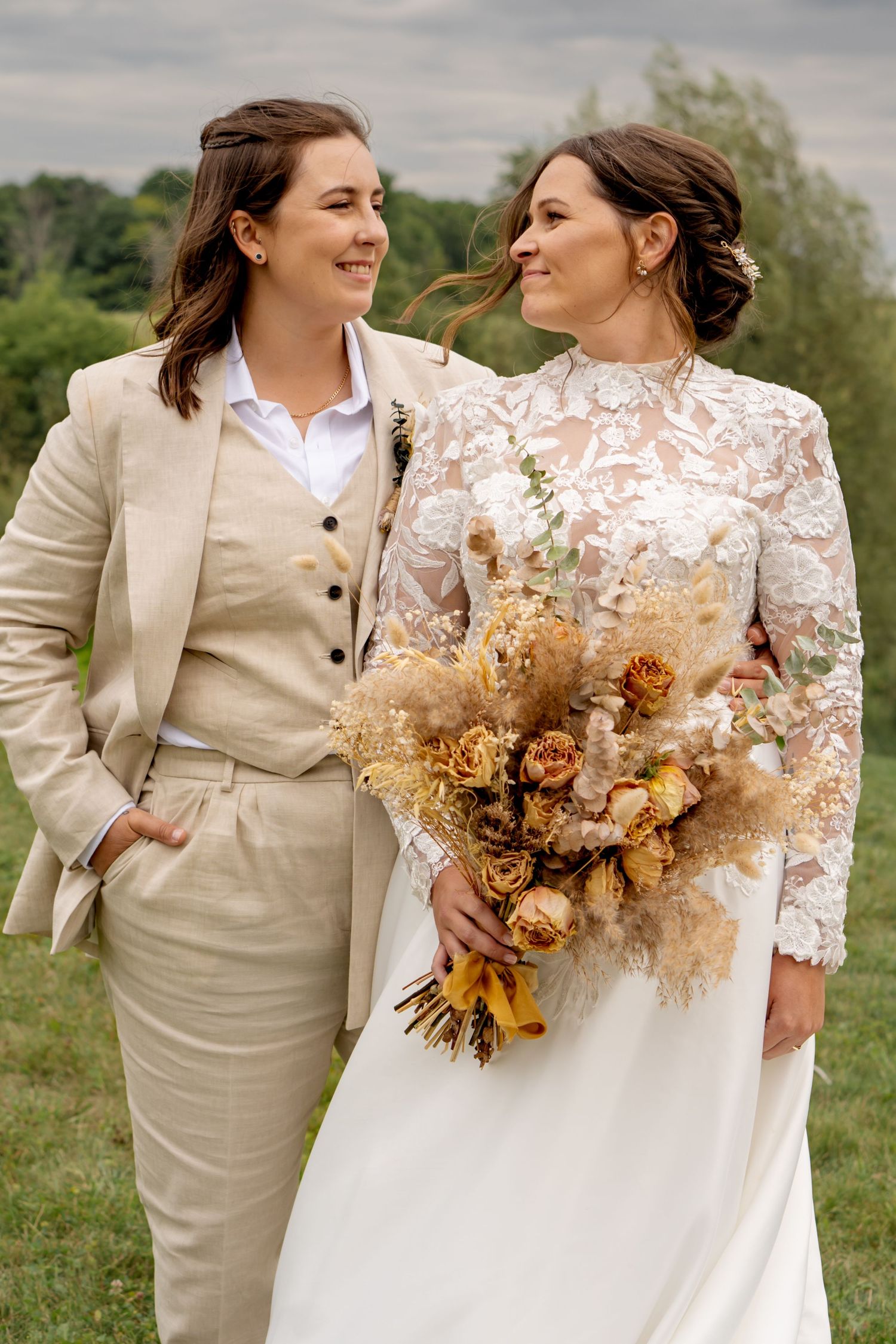 Wedding couple in beige suit and lace dress holding dried flower bouquet in outdoor setting.