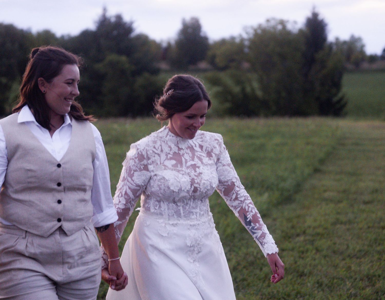 A couple walks hand in hand through a grassy field during a wedding celebration.