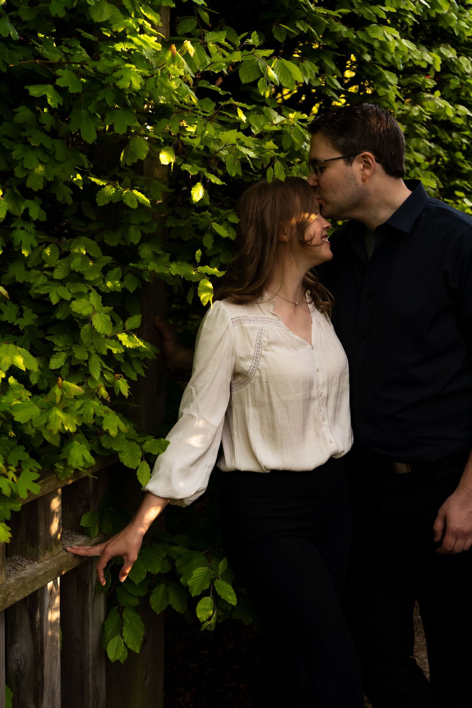 A couple shares a romantic moment under green leafy trees in a garden setting.