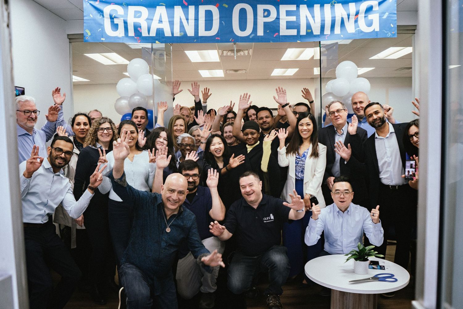 A large group celebrates at an office grand opening event with a blue banner and people cheering.