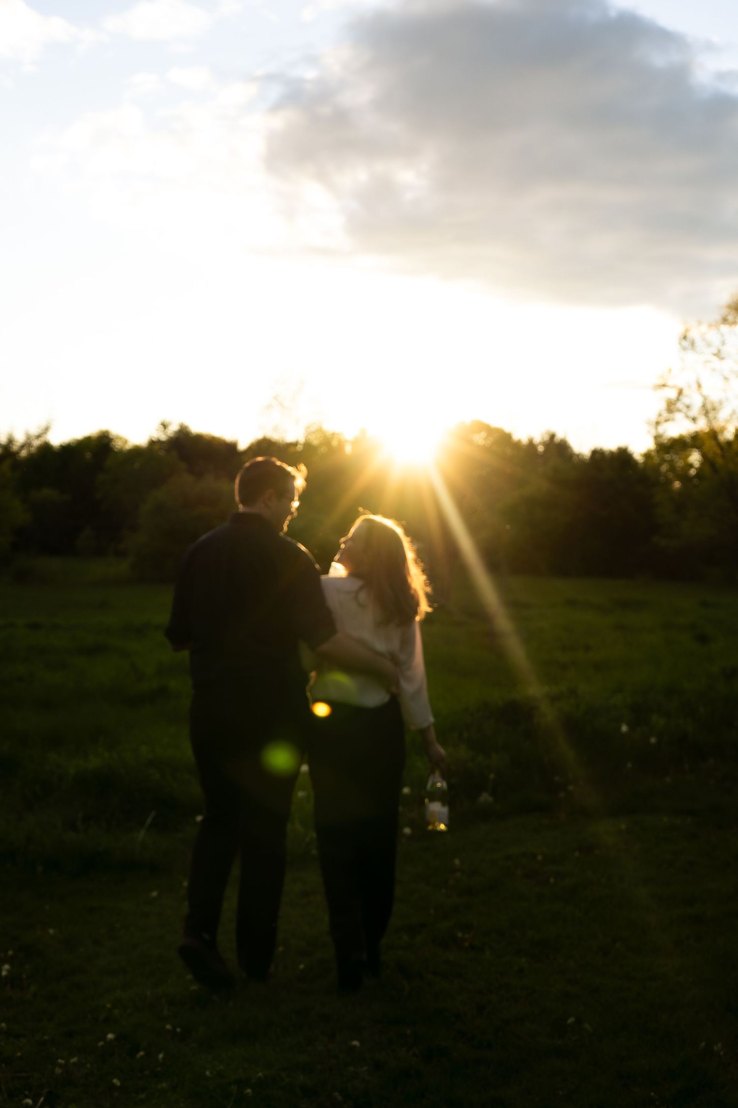 Silhouetted figures stand together in a field at sunset with dramatic sunburst through trees in background.