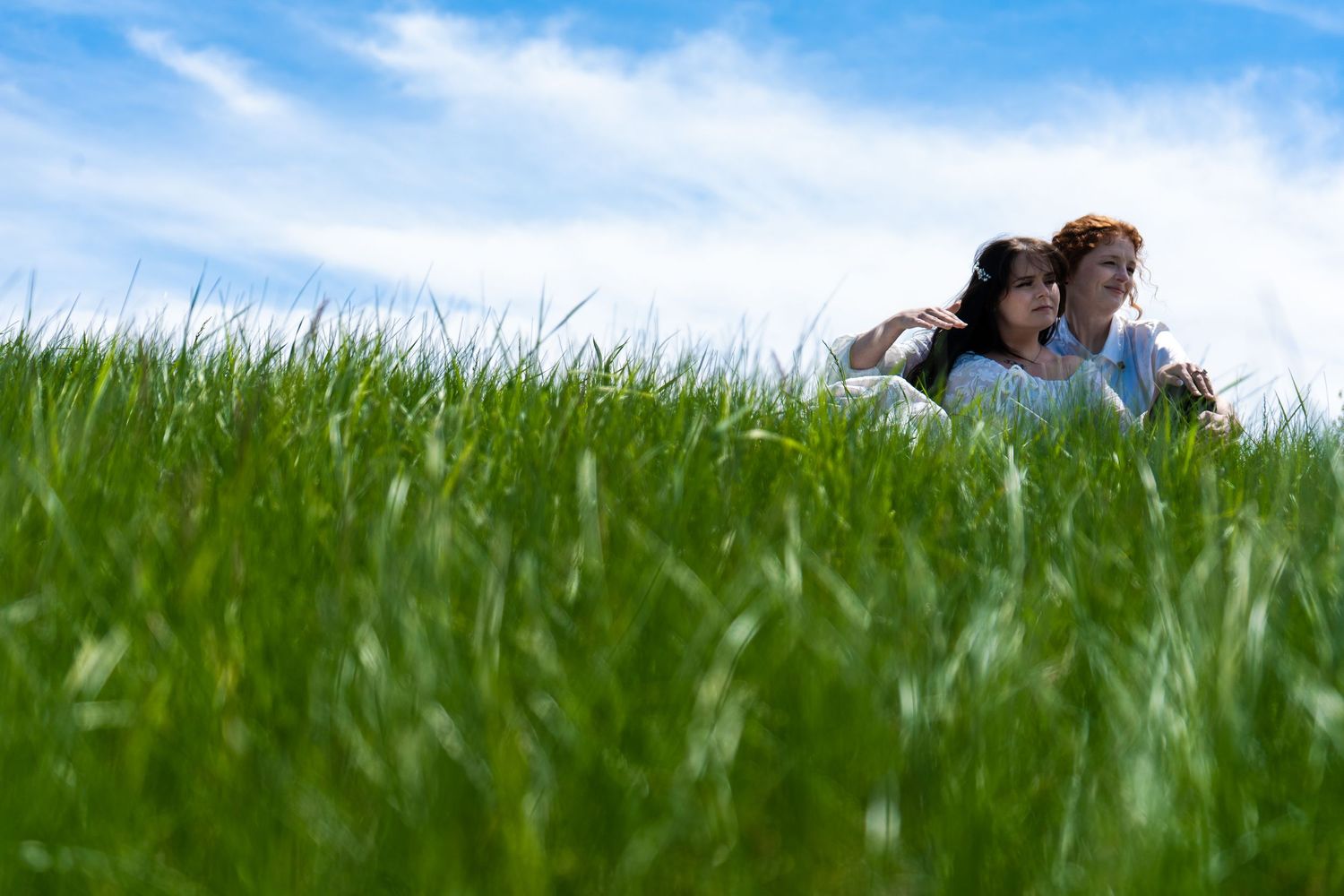 couple sit together in grass field with blue sky.