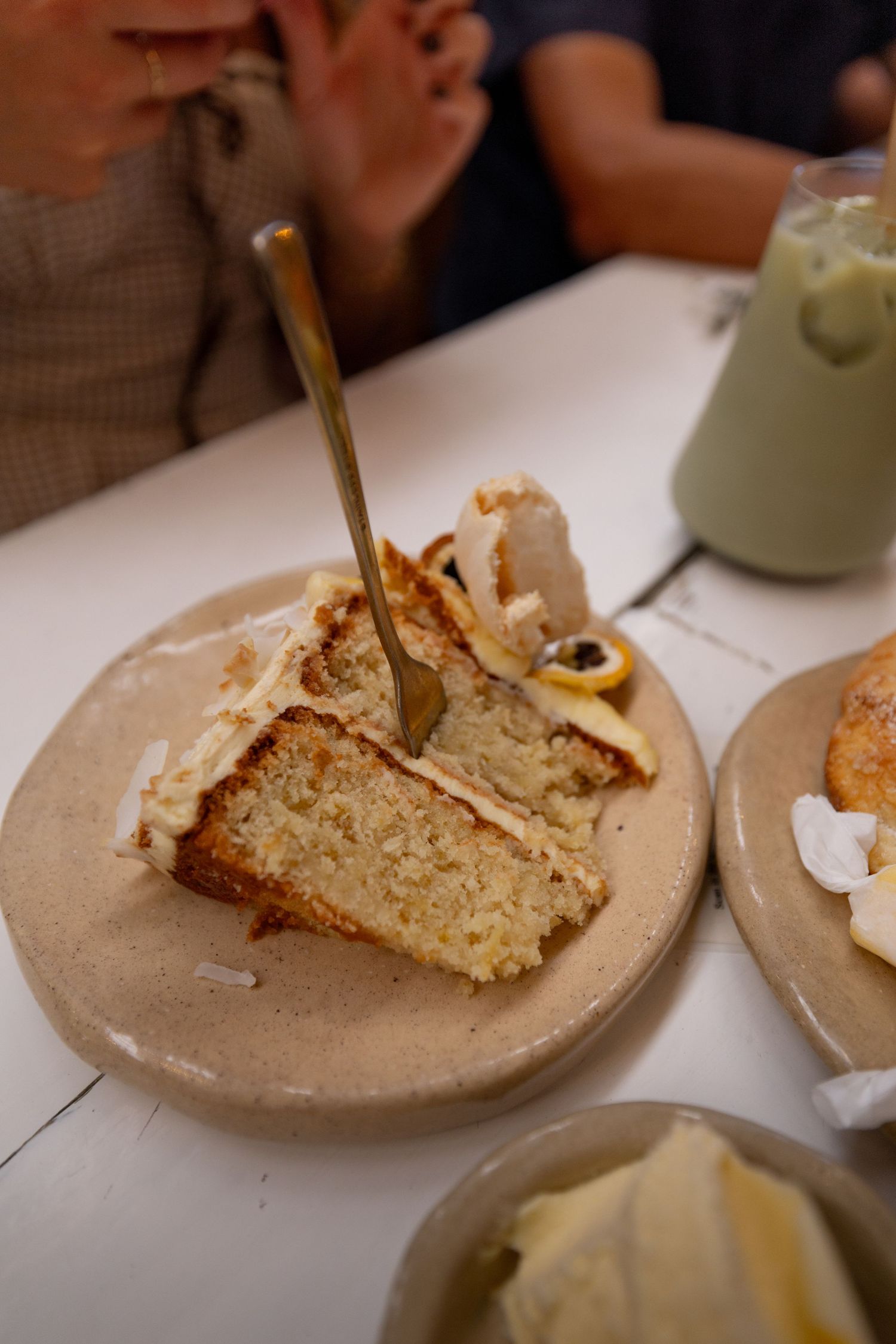 A slice of moist lemon cake with cream frosting on a ceramic plate, served with a fork at a cafe table.