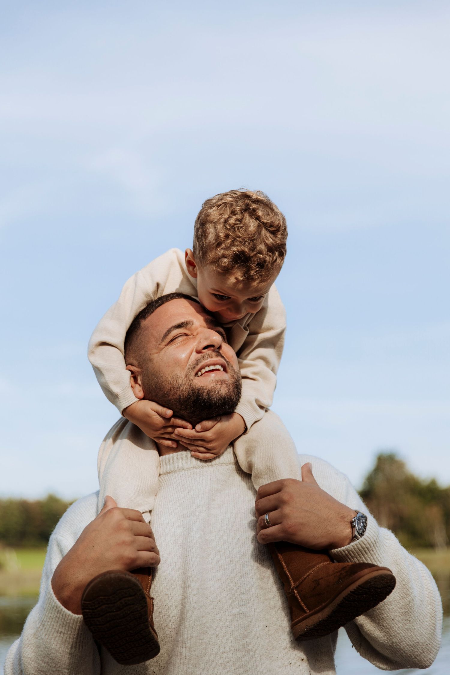 Laughing father carrying his curly-haired toddler son on his shoulders against a blue sky.