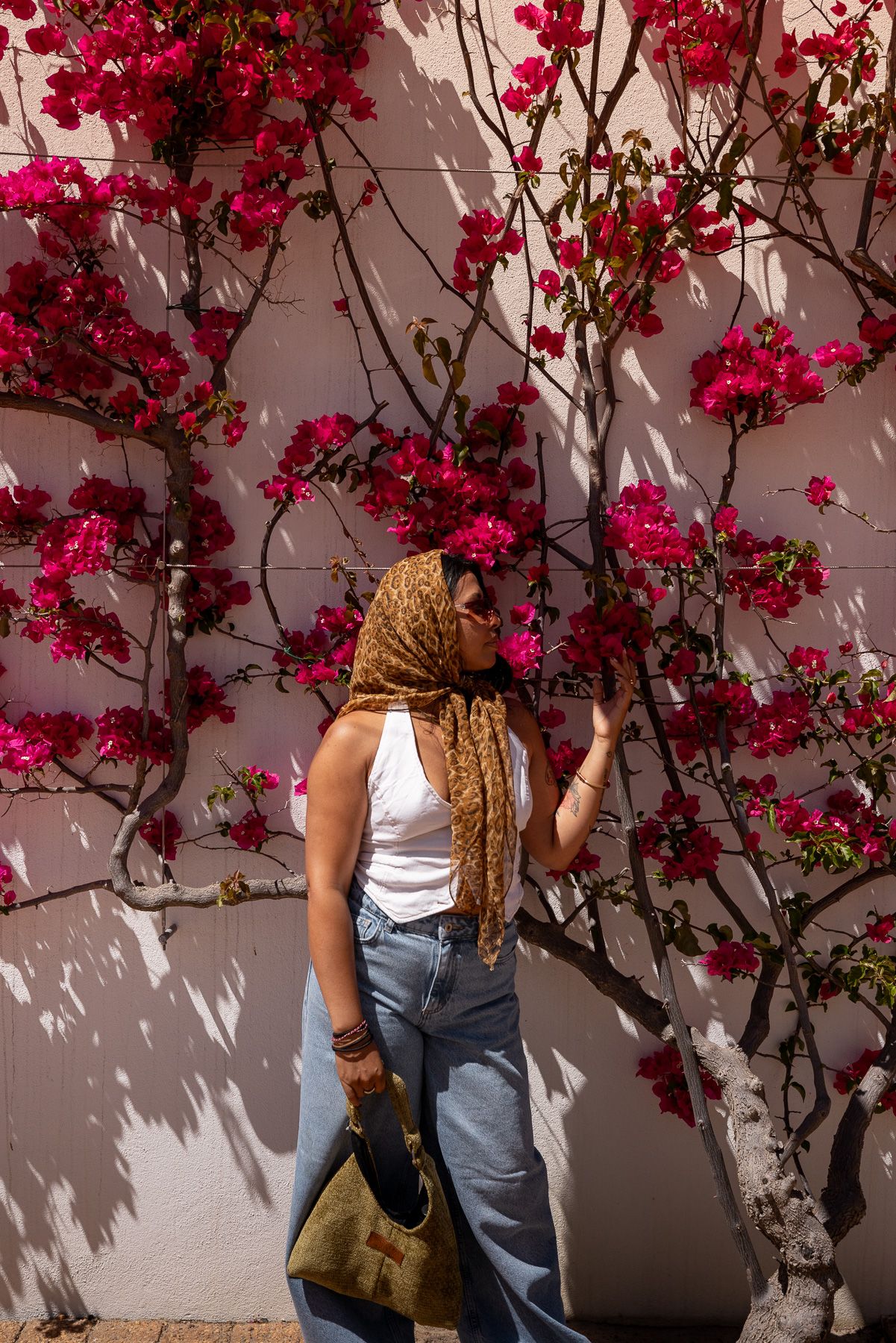 Stylish woman in headscarf and jeans standing by white wall with blooming bougainvillea tree.