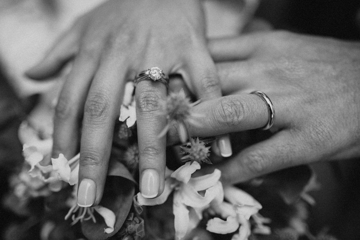 Black and white photo of newlyweds hands with wedding rings resting on a bouquet of white flowers.