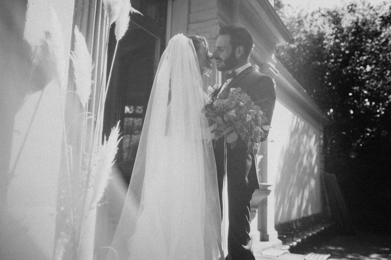 Black and white photo of a bride in a veil and groom in a suit sharing a romantic moment outdoors on their wedding day.
