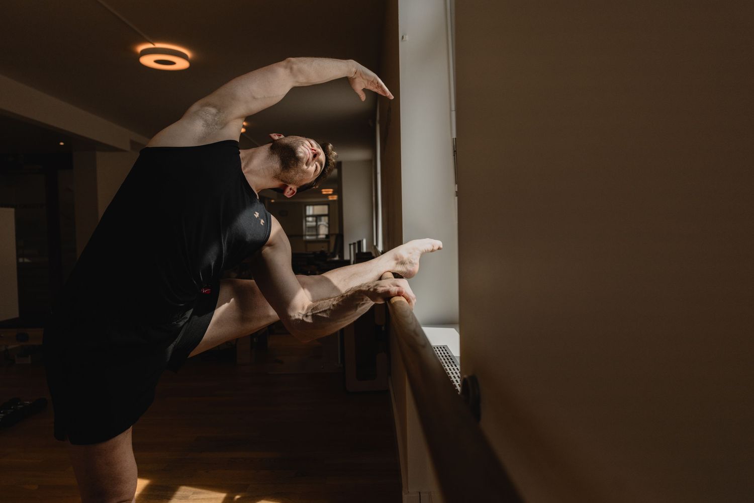 A dancer stretches at a ballet barre in a warmly lit room, wearing a black outfit.