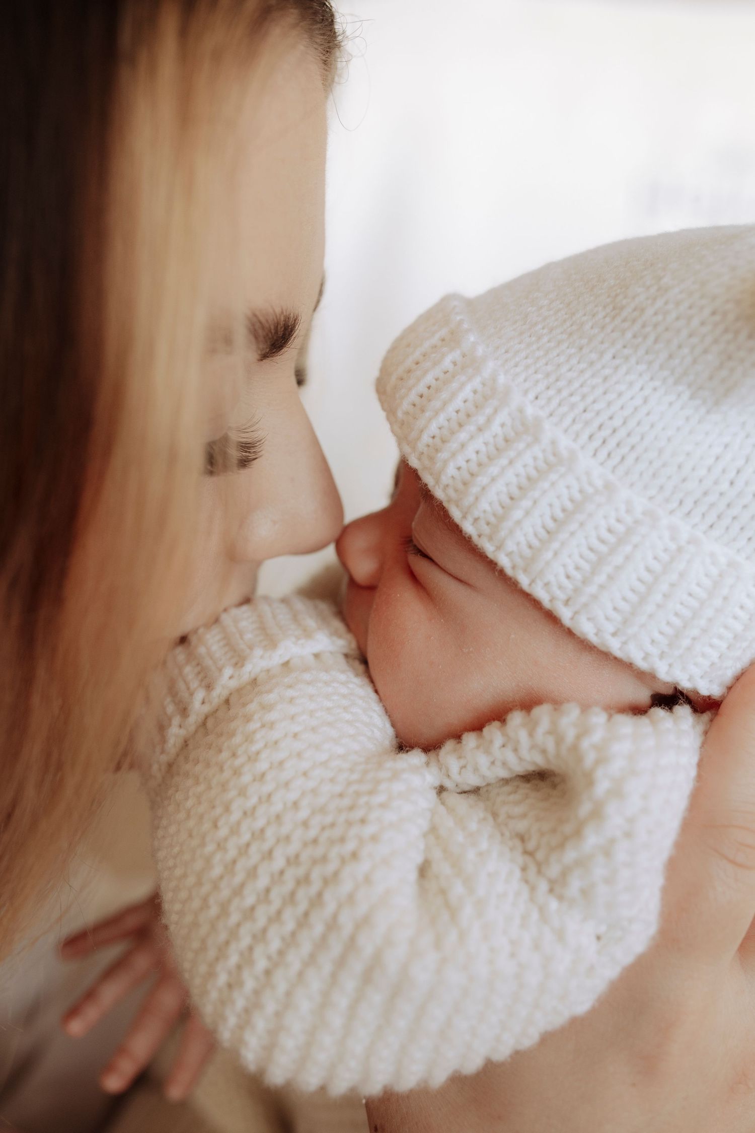 Mother tenderly kissing her sleeping newborn baby wearing a white knit hat and sweater outfit.