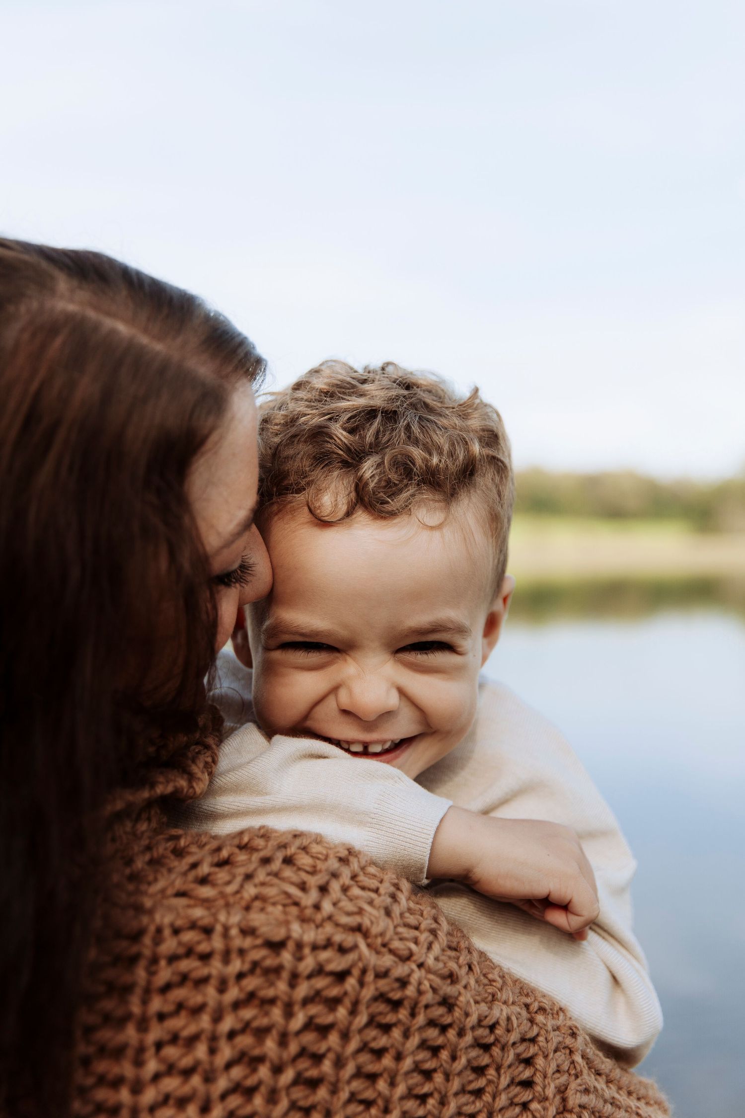 Joyful curly-haired toddler smiling broadly while being held by his mother near a lake.