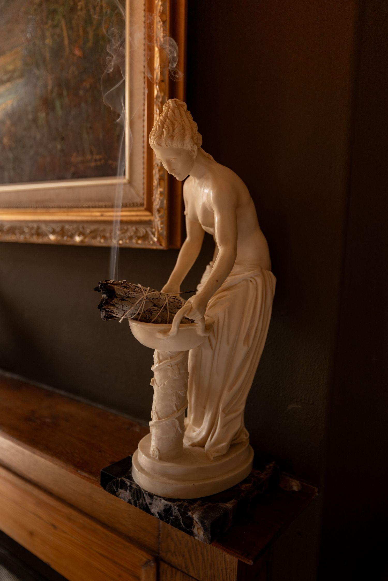 Elegant white marble statue of a classical woman leaning over a basin, displayed on a wooden mantle beside a gold-framed painting.