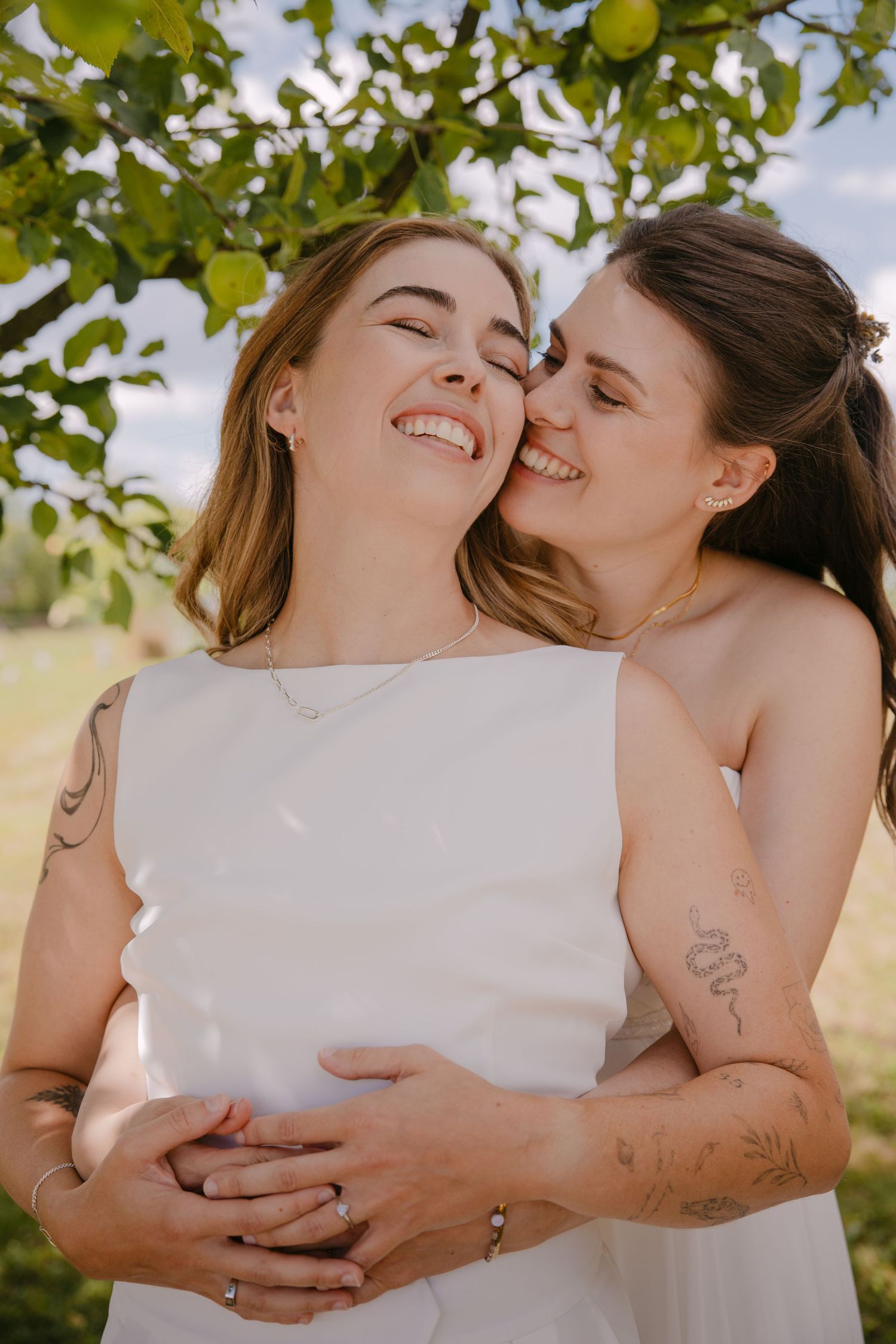 Two brides in white dresses embrace and share a tender moment outdoors under a green tree on their wedding day.